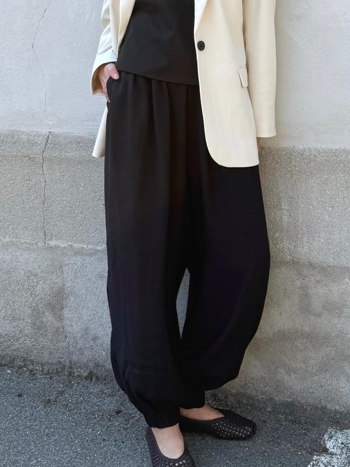 En person iført sorte bukser fra Copenhagen Muse Tenley, en sort top, en cremefarvet blazer og flade sko står ved en væg med struktur.