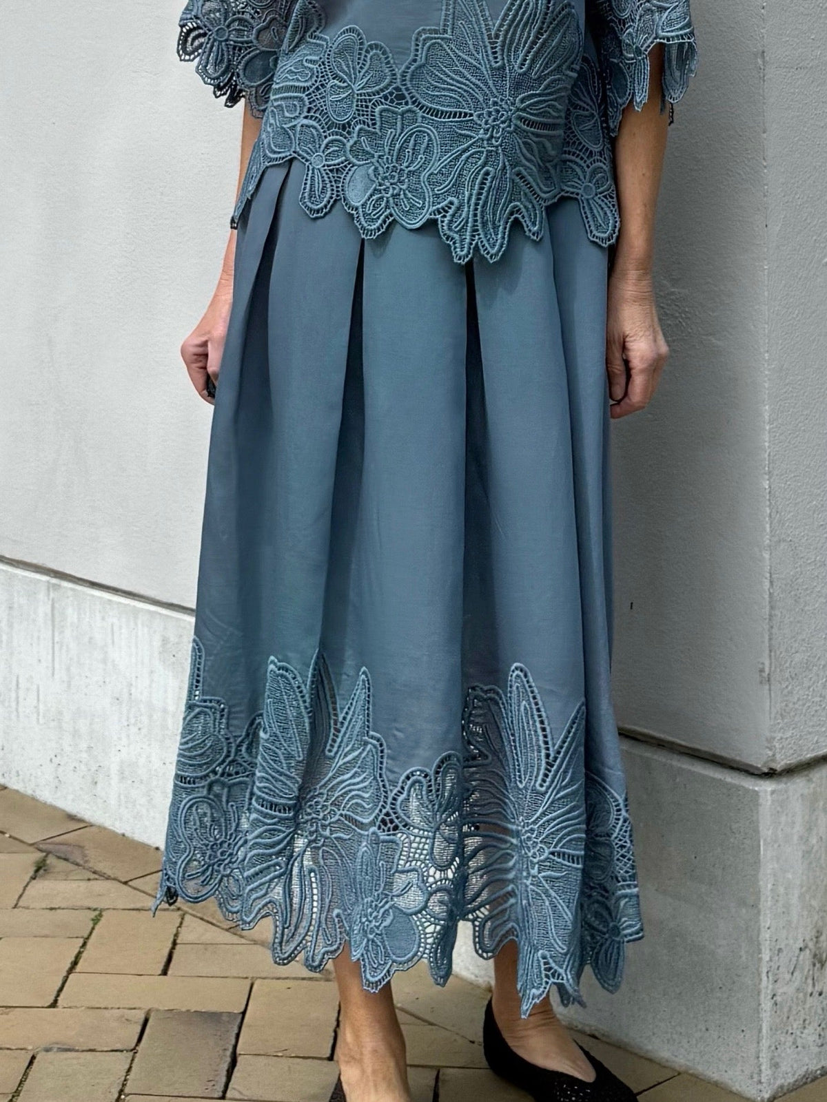En person iført en „Copenhagen Muse – Molly Petrol“-kjole med blomsterbroderi, stående på en flisebelagt fortov.