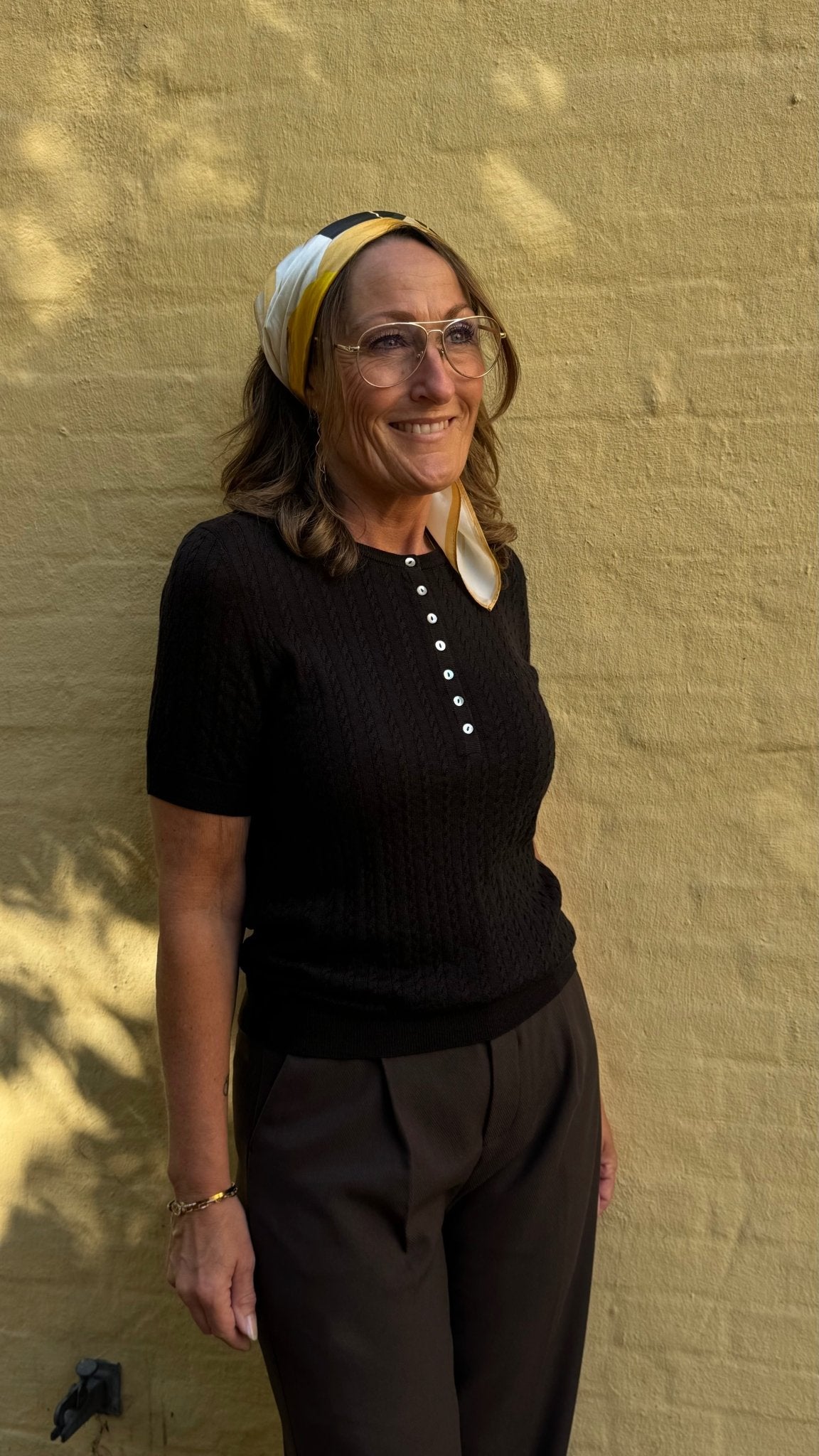 En kvinde med briller, tørklæde, en top fra Copenhagen Muse Eadie Kaffe og brune bukser smiler foran en gul væg med struktur.