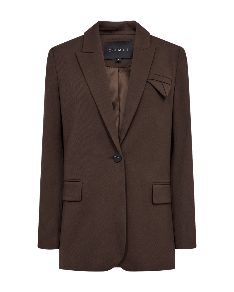 Copenhagen Muse’s Tailor Mørkebrun-blazer har skrå revers, lommer med klap og en elegant foldet detalje på brystet.