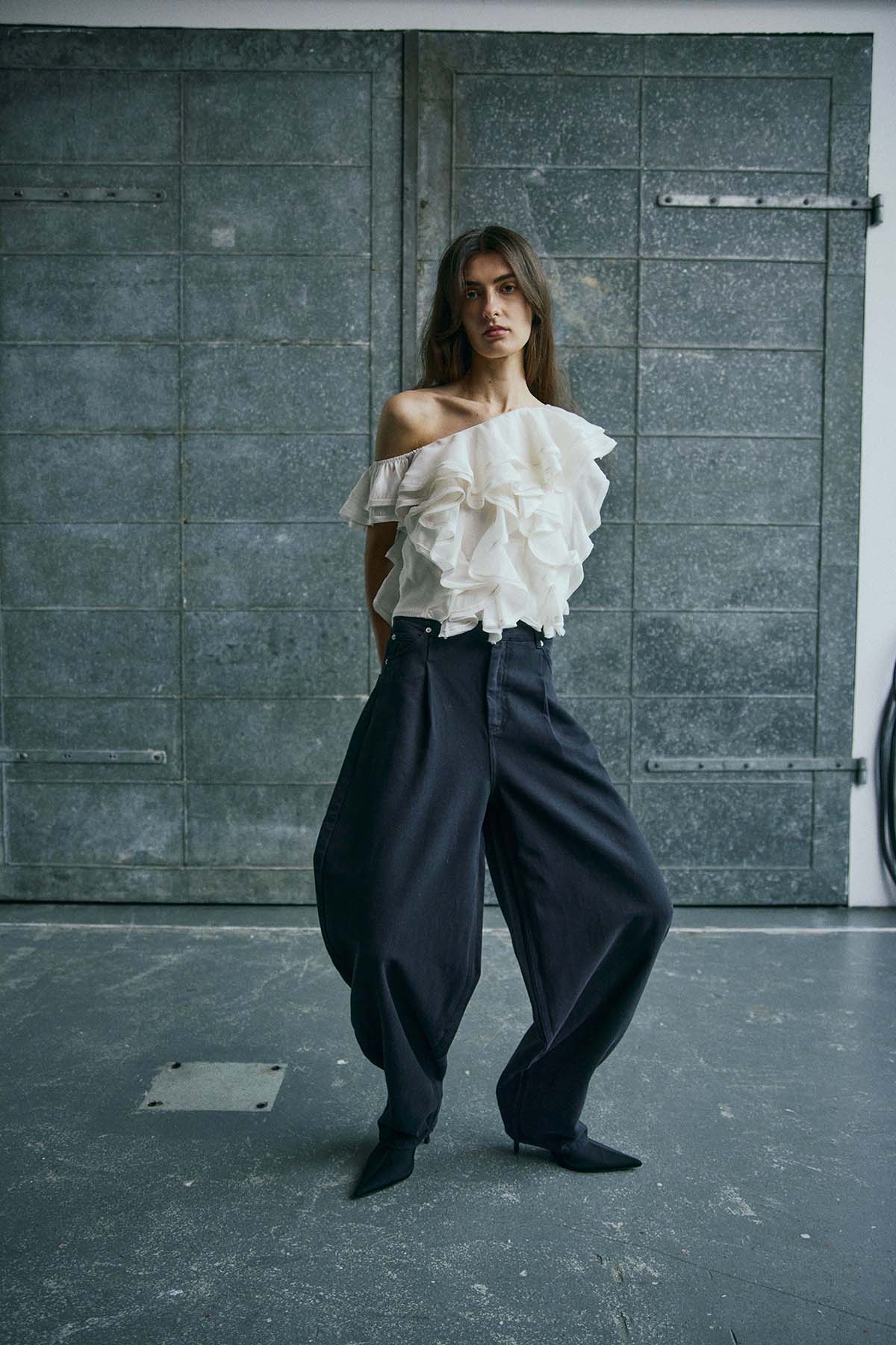 En kvinde står indendørs ved en metaldør iført en Muse Tilly Off White-top fra København og sorte bukser med brede ben.