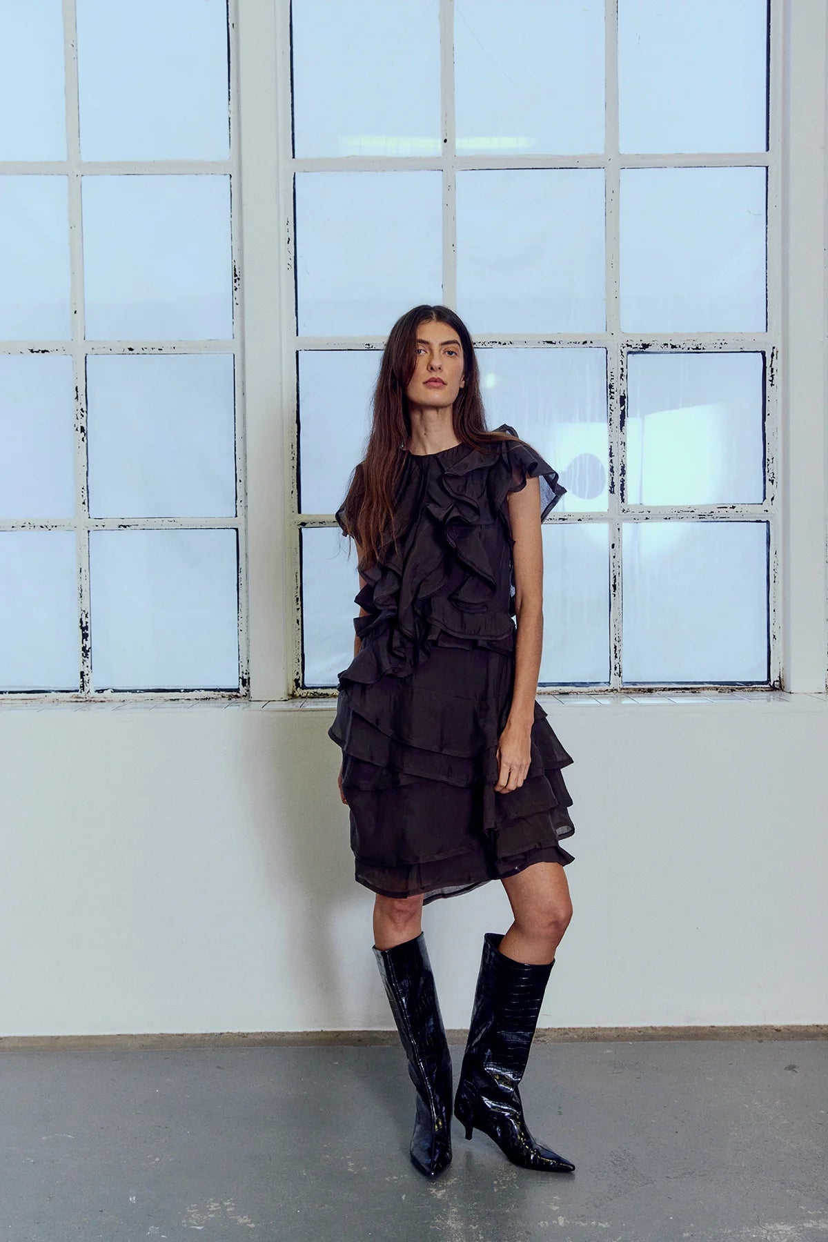 En kvinde i Tilly Mørkebrun-kjolen fra Copenhagen Muse og sorte støvler, inde ved store vinduer.