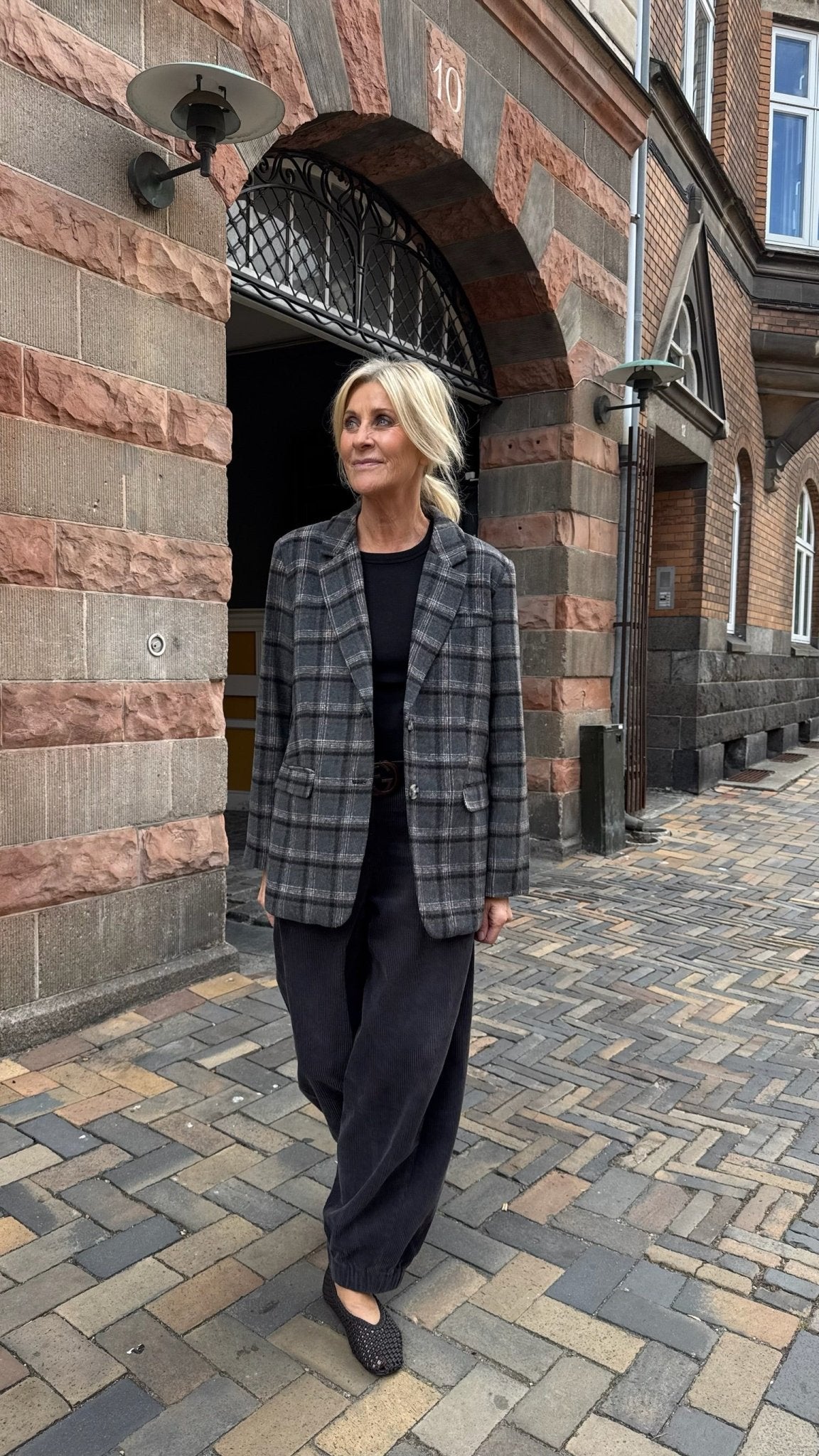 En kvinde iført en Freequent Zango Mørkegrå blazer og mørke bukser går på en brostensbelagt gade tæt på en murstensbygning.