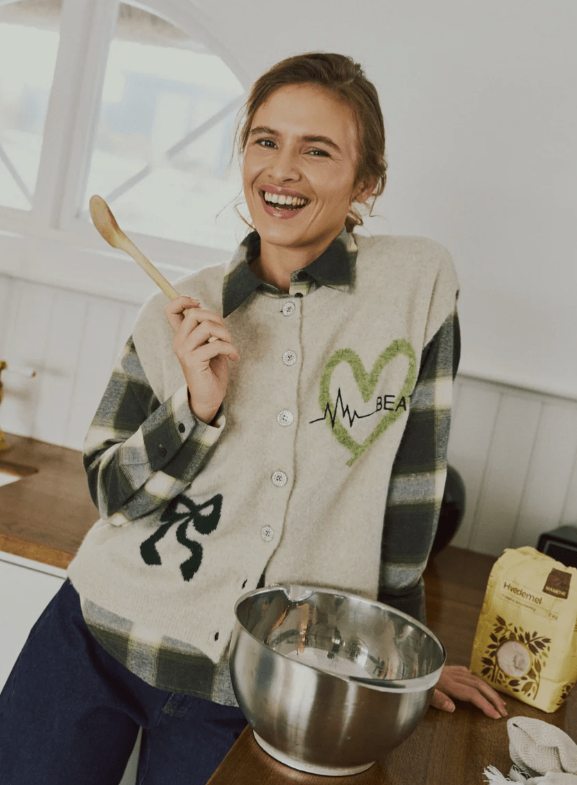En smilende kvinde iført en beige Freequent Nomas-vest, en ternet skjorte og en trøje står i køkkenet ved siden af en røreskål og holder en ske i hånden.