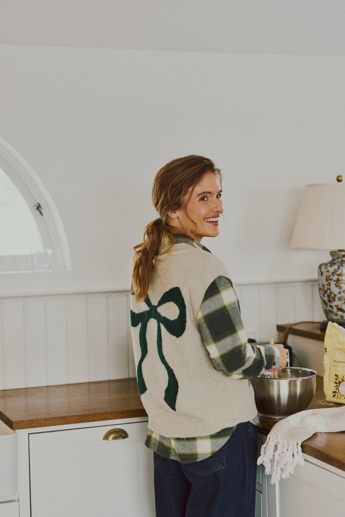 En kvinde i ternet tøj og en beige Freequent Nomas-vest smiler, mens hun bager i et hyggeligt køkken i farven Oyster Grey.