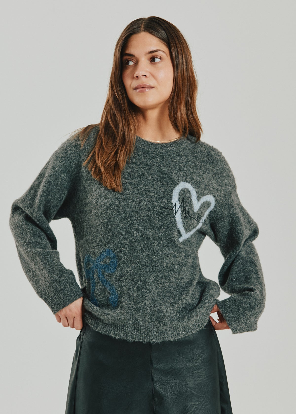 Kvinde iført en Freequent 155794 Nomas Grå sweater med hjerte og sløjfer, kombineret med en mørk nederdel, på en ensfarvet baggrund.
