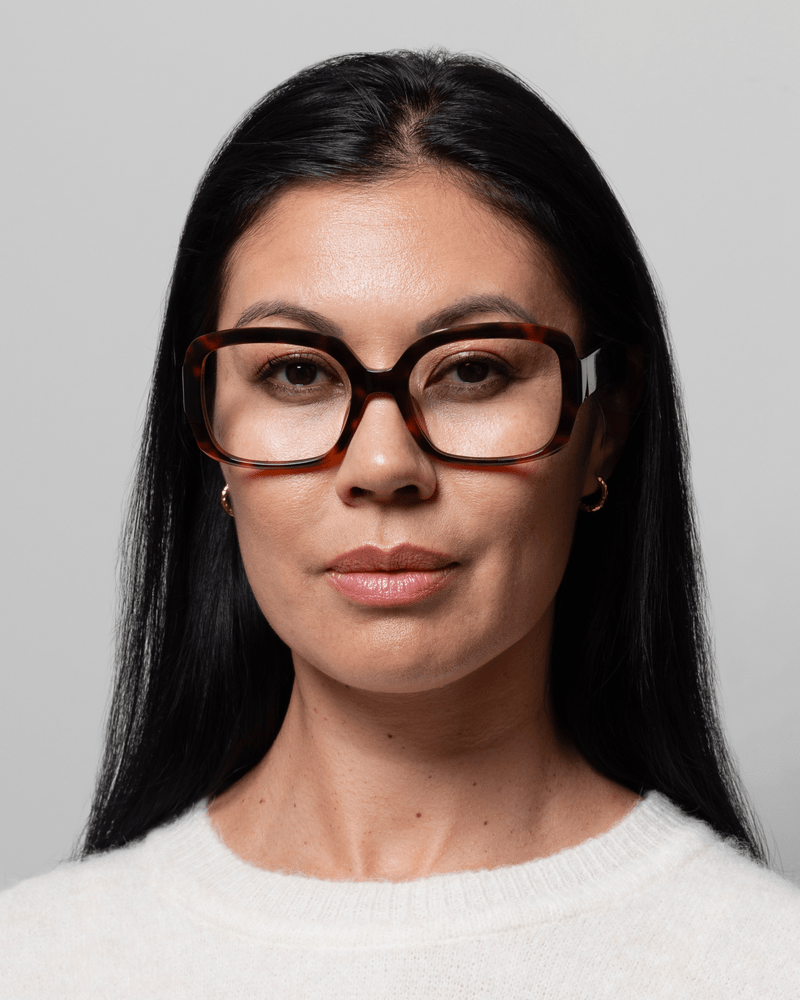 En kvinde med langt sort hår bærer GLAS 152275 Isabella Brun-briller med mønster, hvid sweater, ensfarvet baggrund.