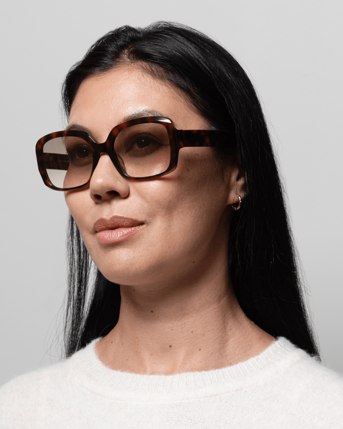 En kvinde med langt, mørkt hår har solbrillerne GLAS Isabella Brun mønstret på og bærer en hvid sweater foran en ensfarvet baggrund.