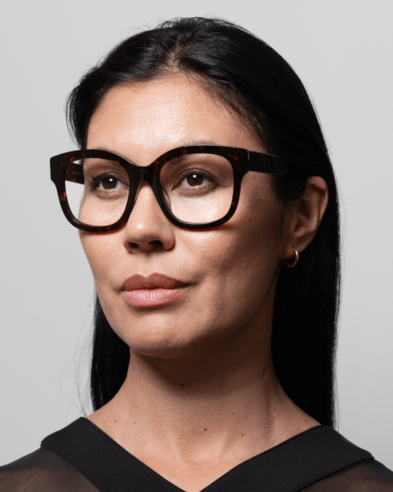 Kvinde med langt mørkt hår i GLAS – briller med Kate Brun-mønster, der kigger til siden foran en ensfarvet baggrund.