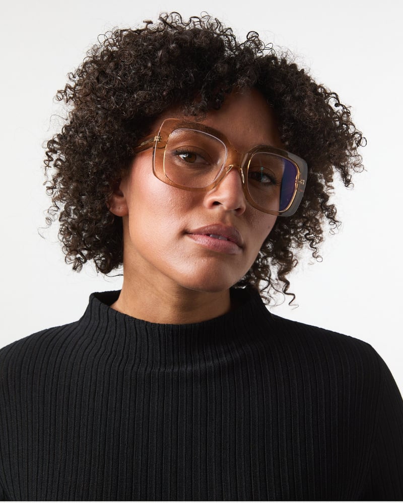 Kvinde med krøllet hår iført GLAS – Mio Camel oversize-briller og sort ribstrikket top, der står med ansigtet vendt fremad foran en hvid baggrund.