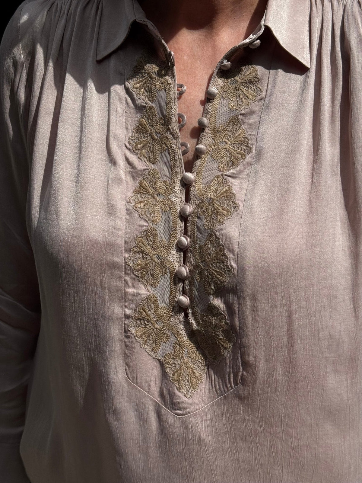 Personen i Gustav Sikka Lysebrun: lyserød bluse med guldbroderi i blomstermotiv og stofbeklædte knapper foran.