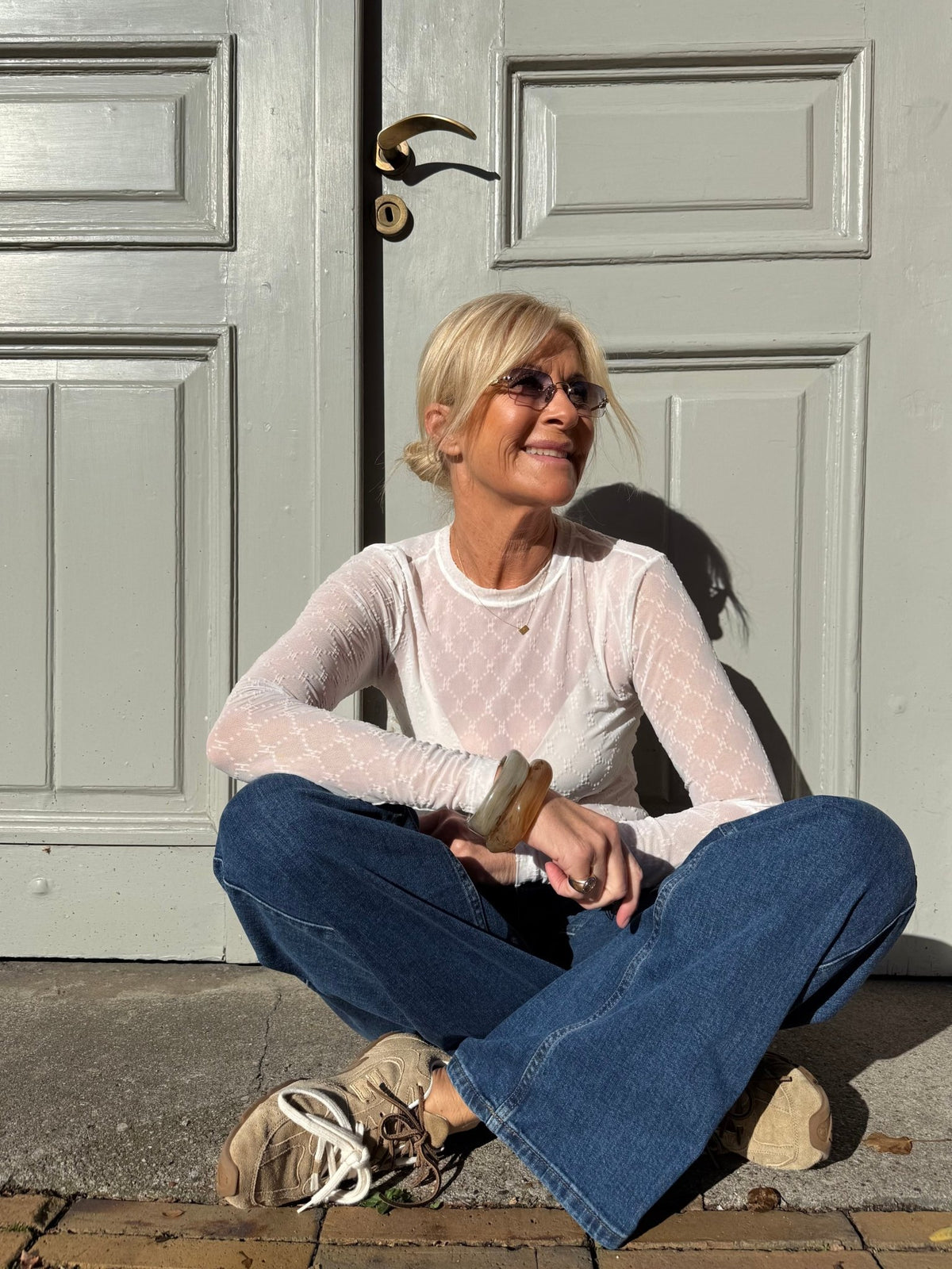 En kvinde i en HYPE THE DETAIL-netbluse i offwhite og solbriller sidder med benene krydsede og smiler i sollyset foran de grå dobbeltdøre.