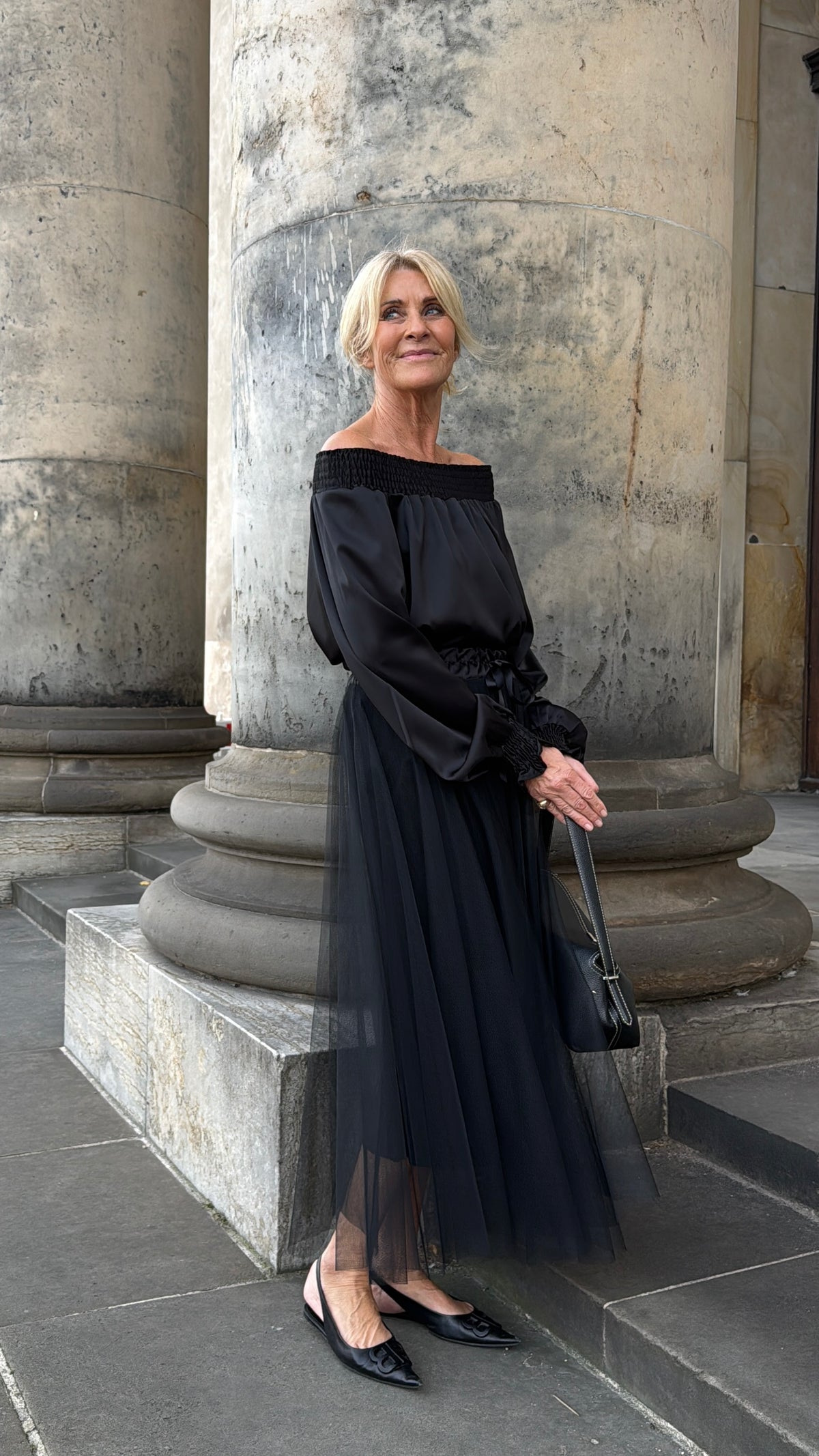 En kvinde i en lang sort kjole står ved de stenkolonner, hvor Marta du Château MDCVerona Sort-tasken hænger, og kigger let opad.