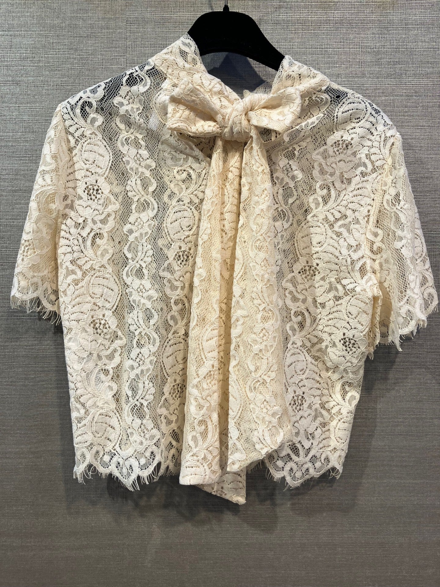 Imperial 154149 | RIT8 Offwhite kortærmet bluse med elfenbensfarvet blonder og sløjfe ved halsen, hængende på en bøjle foran en struktureret baggrund.