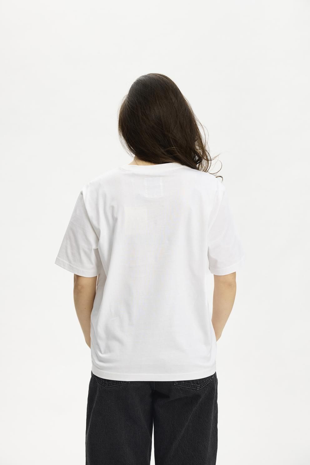 En person med langt, mørkt hår har en Kopenhaken Kenai Lady Hvid T-shirt og sorte bukser på og står med ryggen til kameraet.