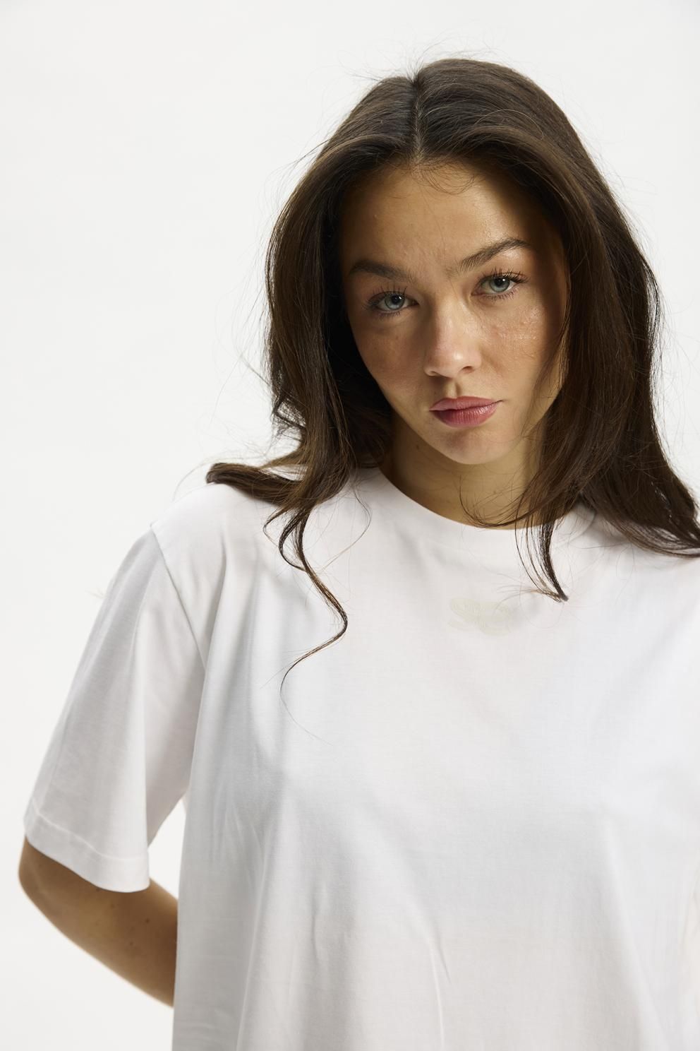 En kvinde med langt brunt hår har en Kopenhaken – Kenai Lady Hvid T-shirt på og står foran en hvid baggrund.