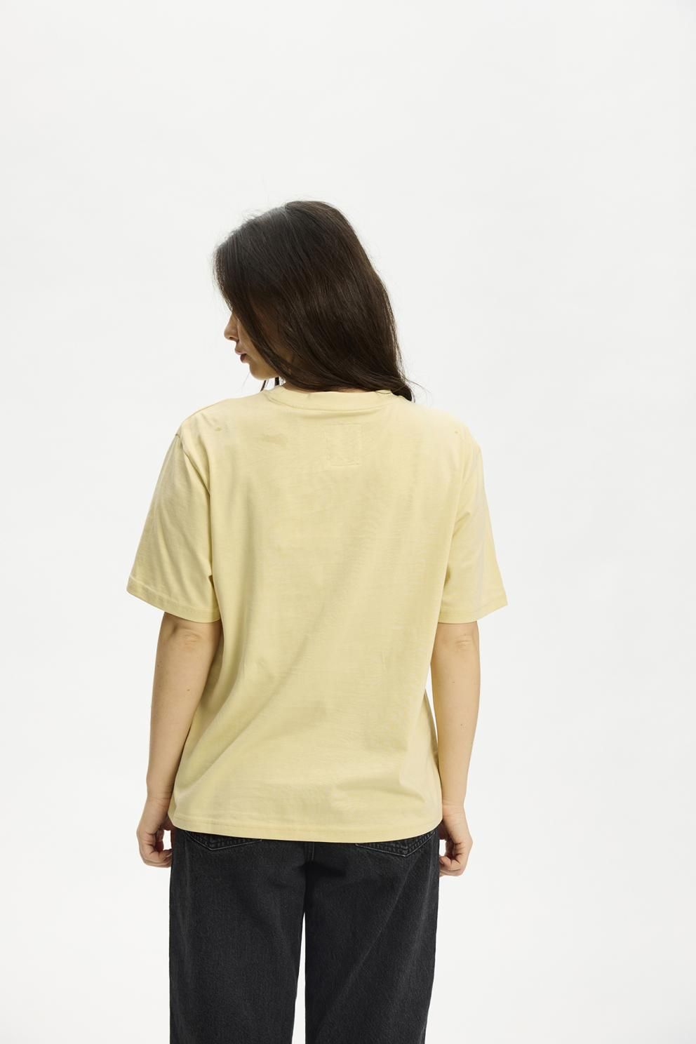 En person, der har den gule Kopenhaken Kenai Lady Gul-T-shirt på, står med ryggen til os, og man kan se hendes lange brune hår.
