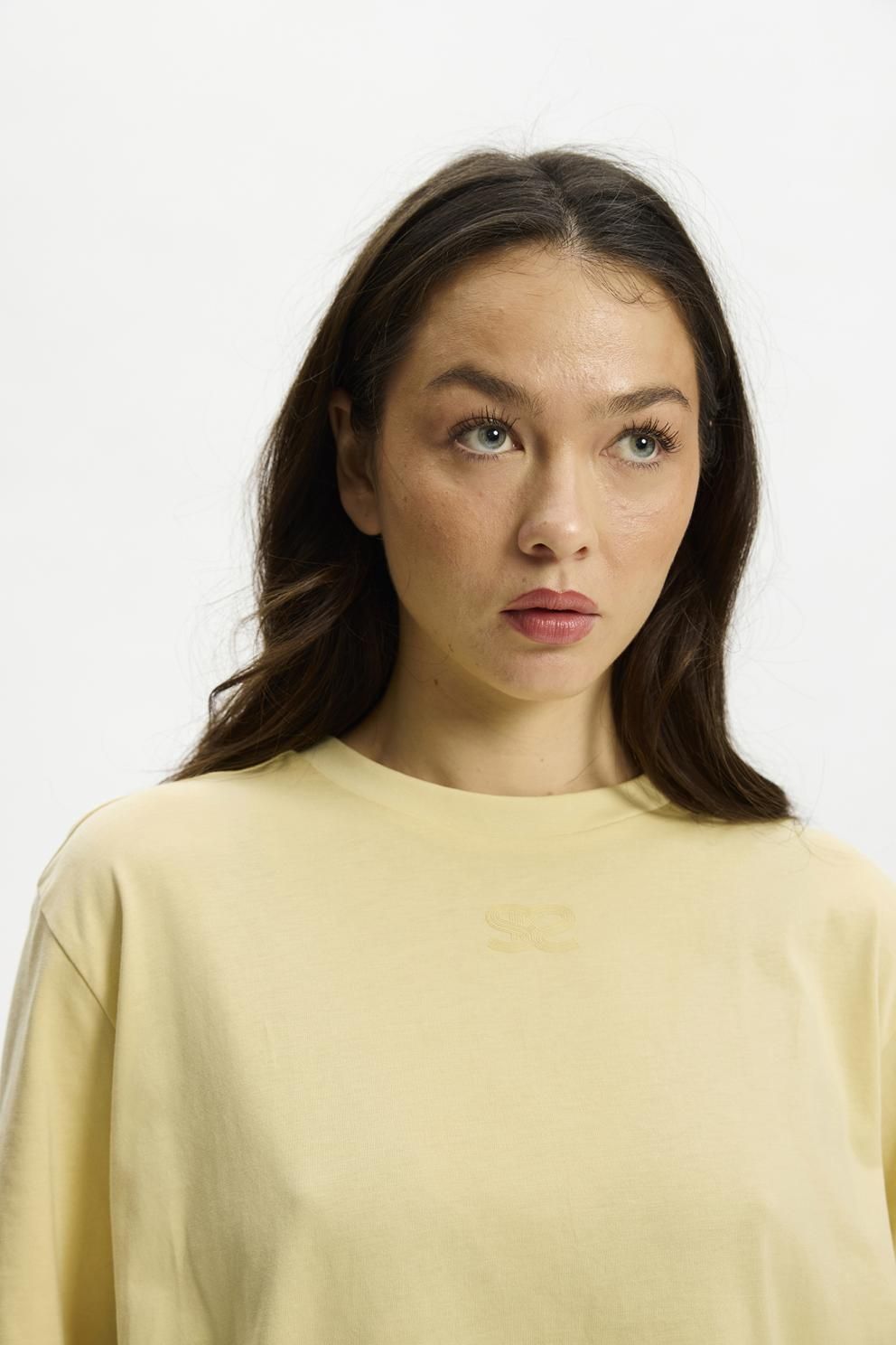 En kvinde med langt brunt hår iført en gul Kopenhaken Kenai Lady Gul-T-shirt, der kigger op mod en hvid baggrund.