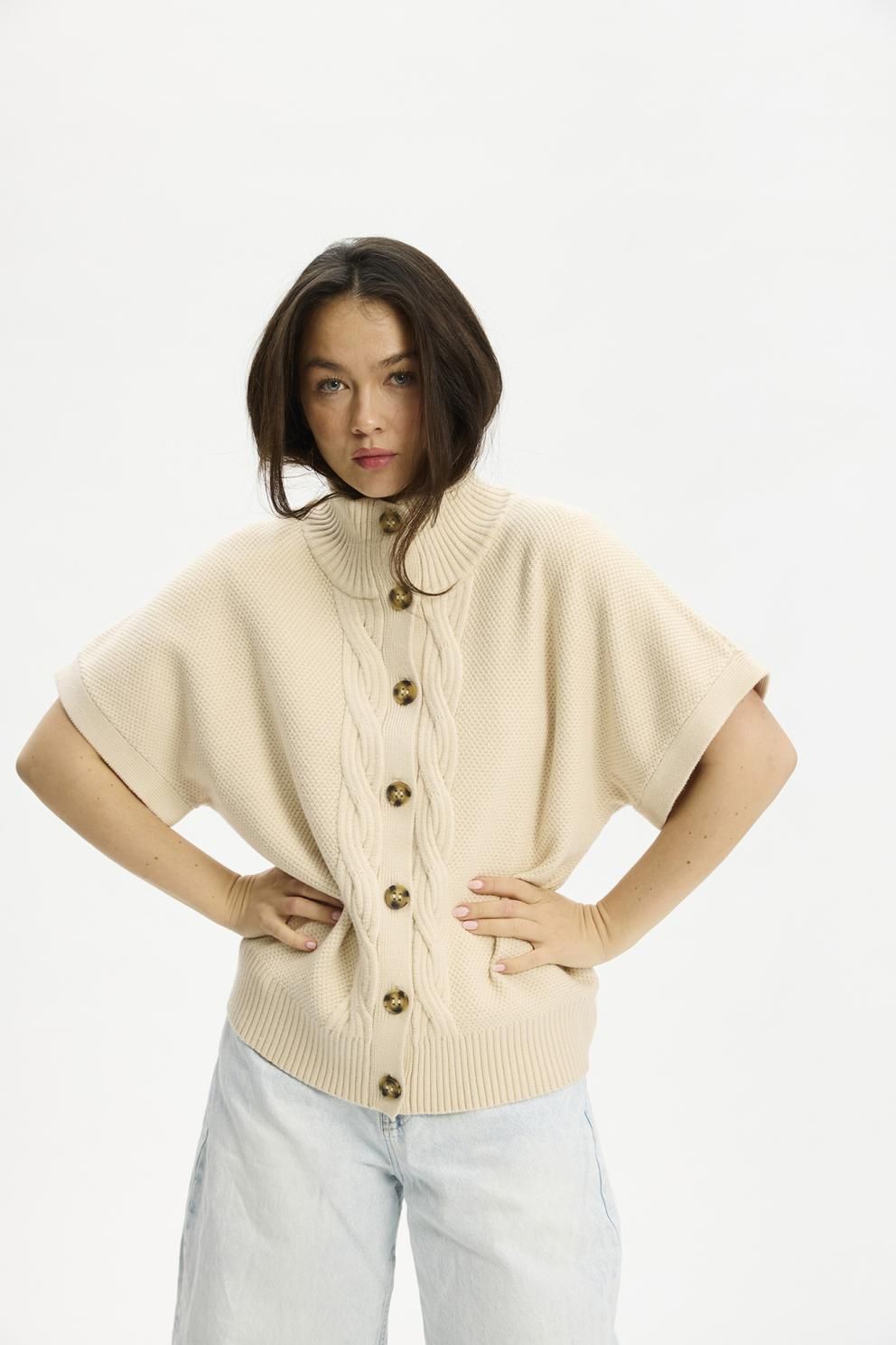 En kvinde iført en Nexus Sand-cardigan fra Kopenhaken og lyseblå jeans står foran en hvid baggrund.