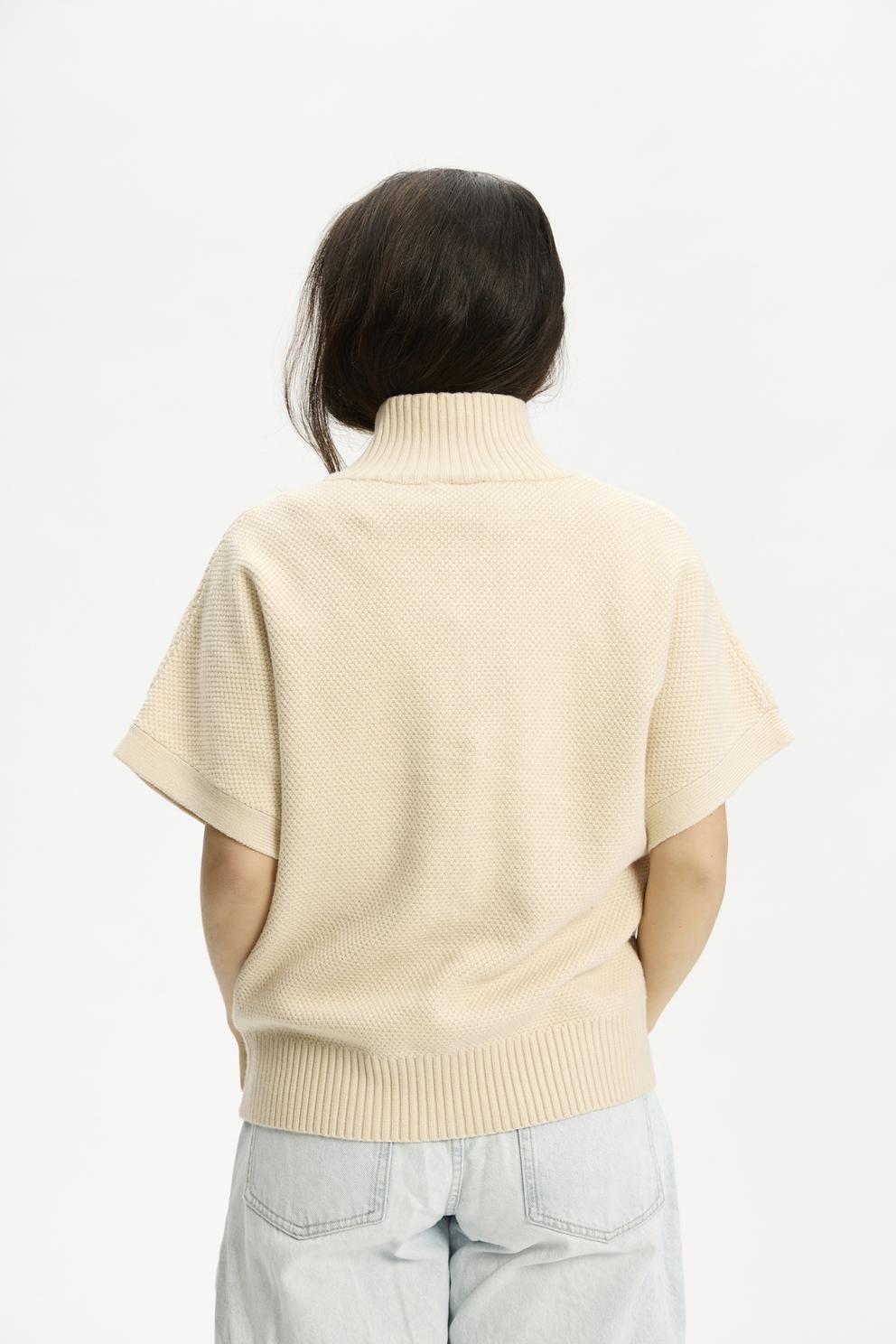 Bagfra set: Kopenhaken Nexus Sand – en strikket rullekravesweater med kort ærme og struktureret strik, stylet med jeans i lys vask.