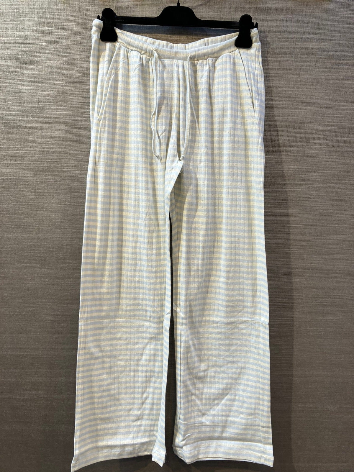 København – Lyseblå pyjamasbukser med bløde striber, perfekte til afslapning, hænger på en sort bøjle foran en grå væg.