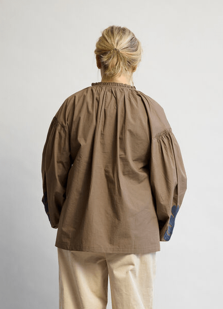 En person med blondt hår sat op i en knold, iført en Kopenhaken Rover Brun-trøje og beige bukser, der står med ryggen til.