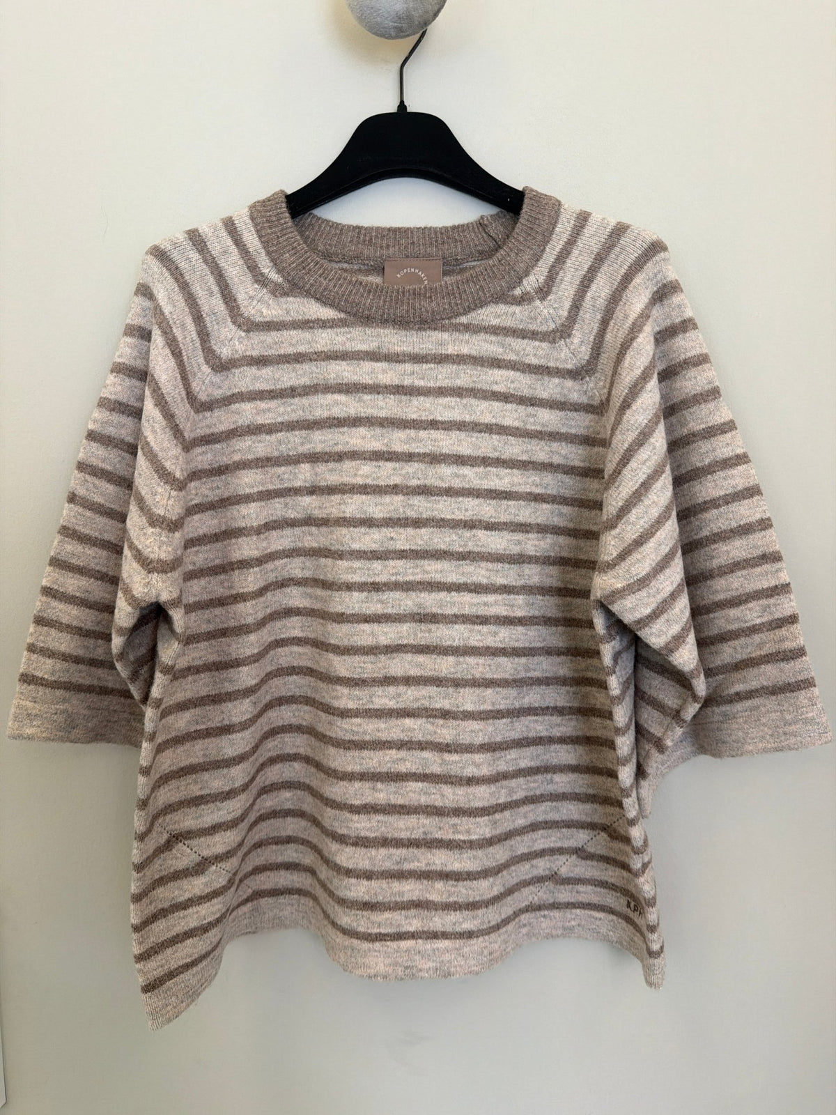 En strikbluse fra Kopenhaken Carla Brun på en sort bøjle, med mønster i beige og taupe, foran en lys væg.