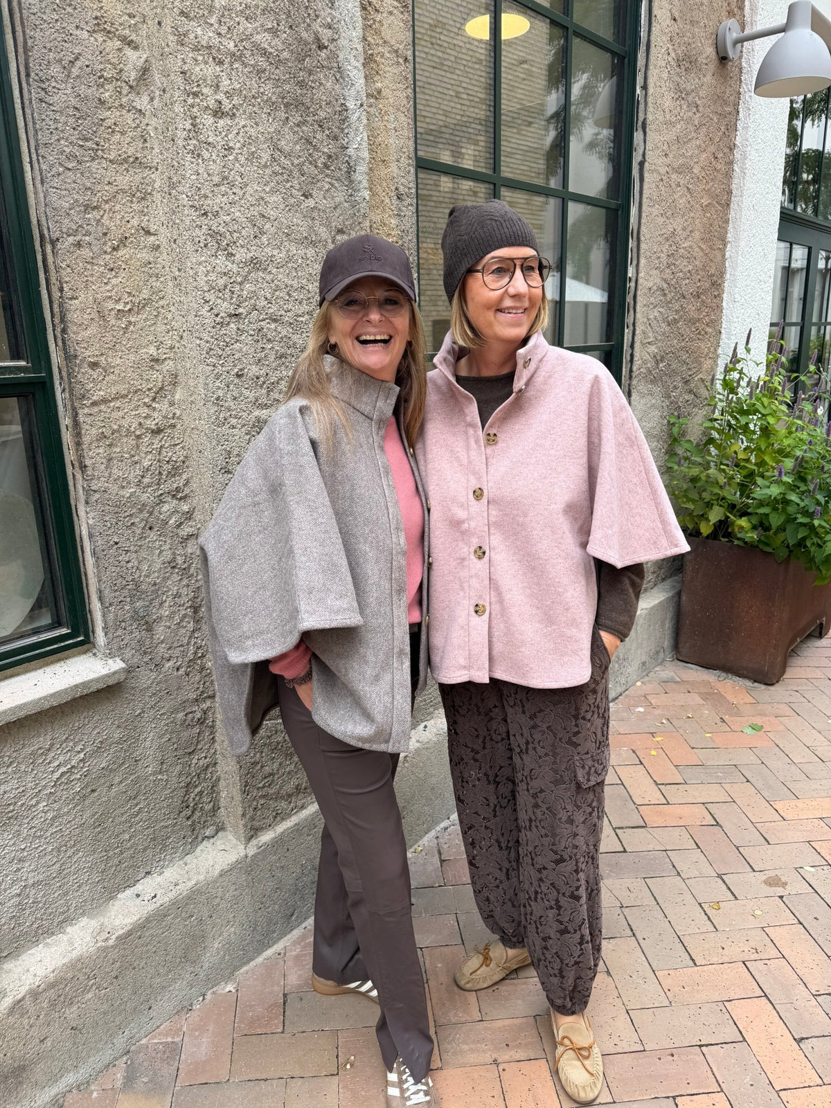 To kvinder i efterårstøj, hvoraf den ene har en Kopenhaken Isla 2 Rosa-poncho på, smiler sammen på en fortov af mursten.