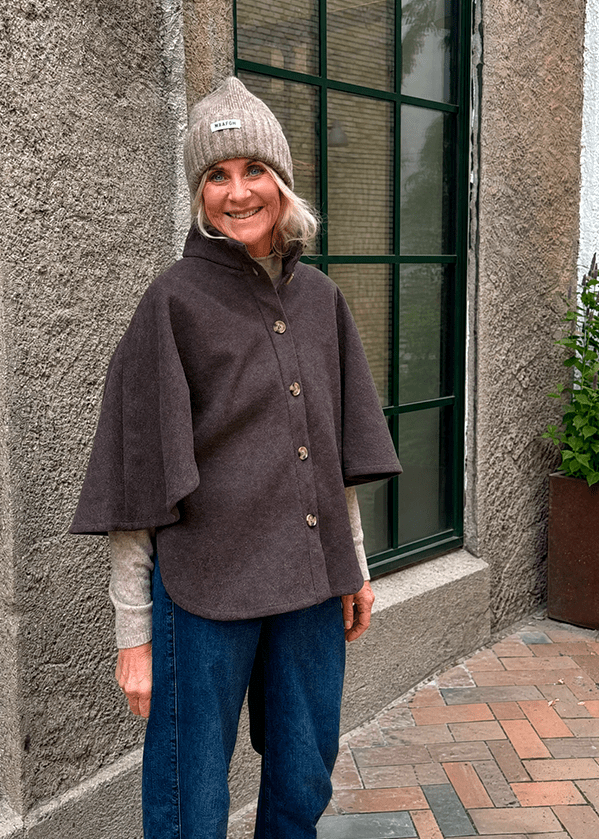 En kvinde iført en Kopenhaken Isla 2 mørkebrun poncho, en beige strikhat og jeans står udendørs ved et stort vindue.