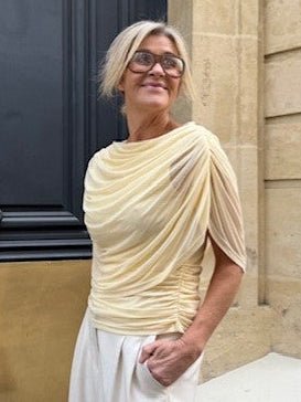 Kvinde med gråt hår iført en Luxzuz-bluse fra Queenie Lysegul og hvide bukser, der står ved en stenmur.