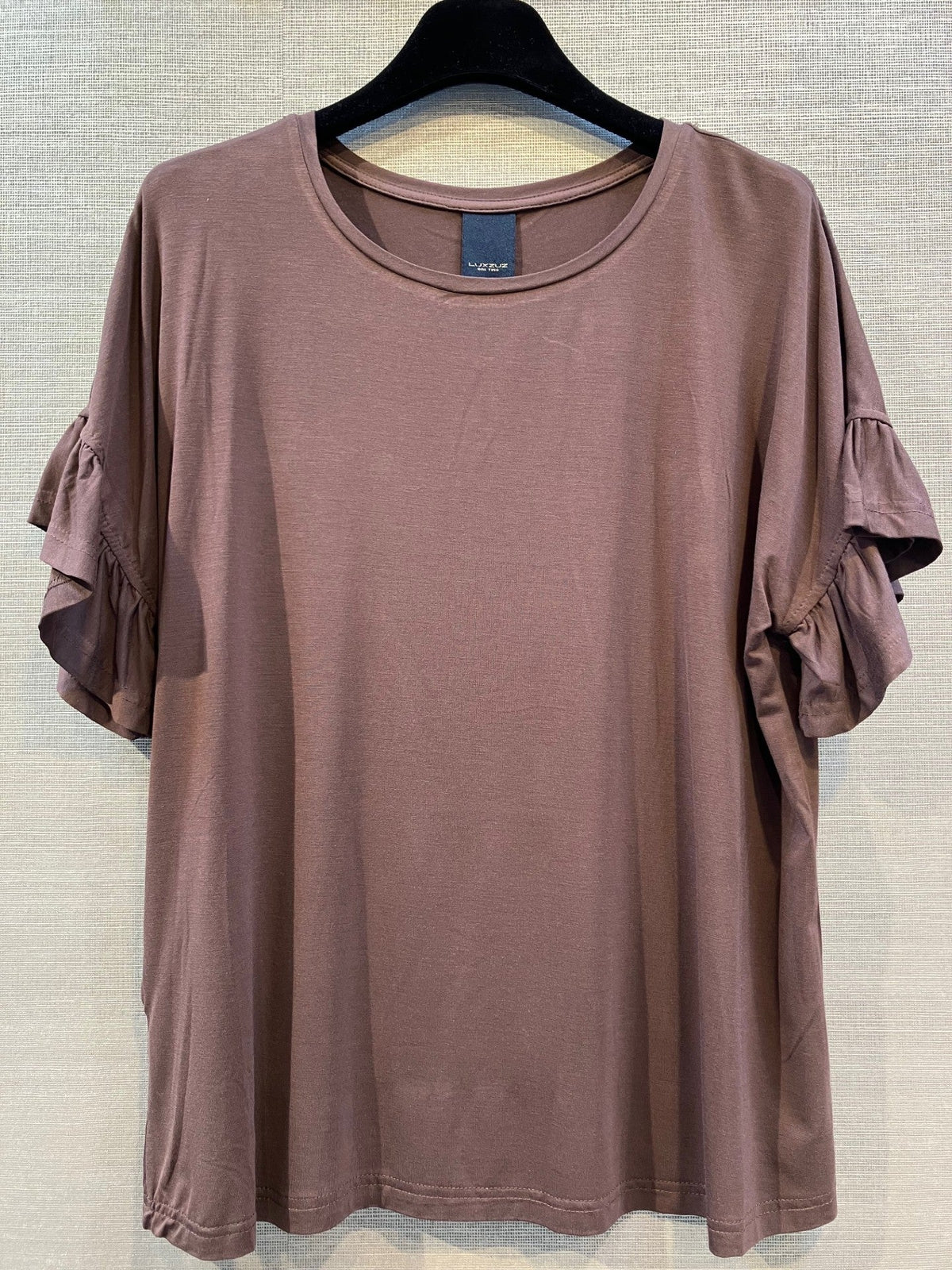Luxzuz Seija mørkebrun T-shirt med flæseærmer, hænger på en sort bøjle foran en neutral, struktureret baggrund.