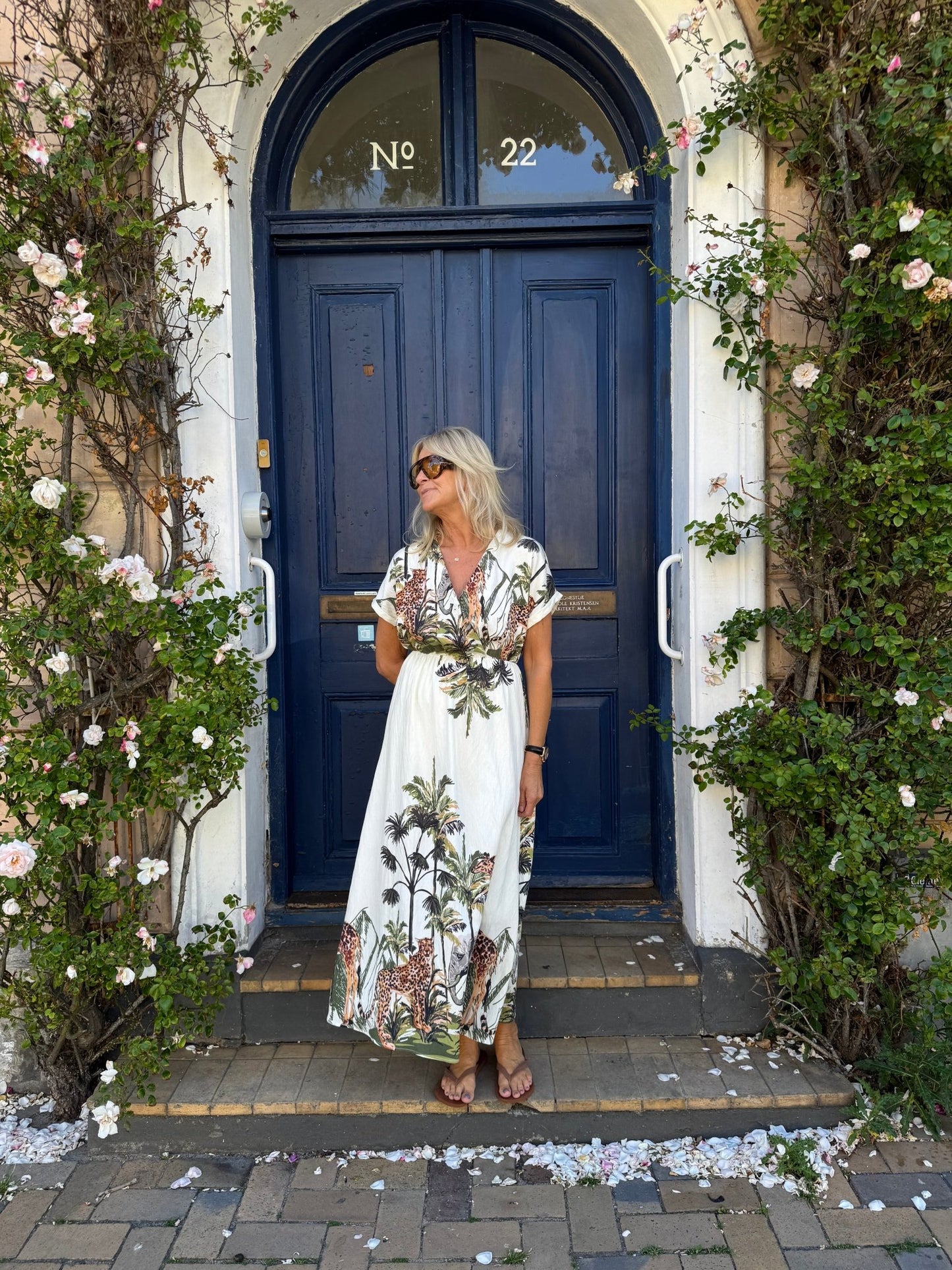 En kvinde i en mønstret kjole fra Luxzuz Via Off-White står foran en blå dør, omgivet af grønne ranker og hvide blomster.
