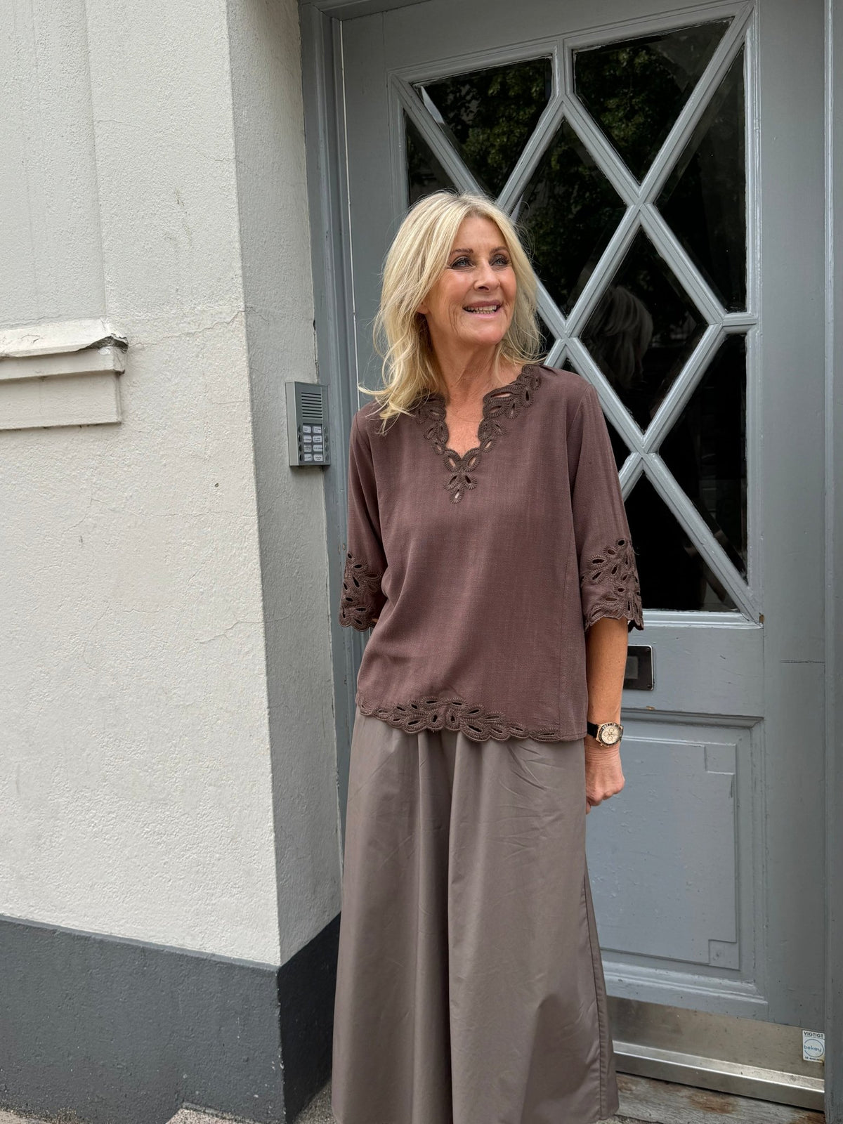 En blond kvinde ved en grå dør har en Luxzuz Vicha Brun-bluse på sammen med brede grå bukser.