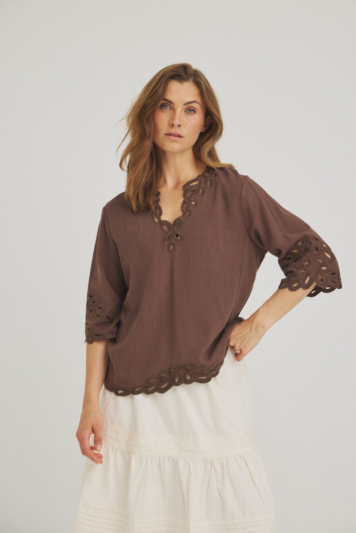 En kvinde poserer iført Luxzuz Vicha Brun, en mørkebrun bluse i broderie anglaise, og en lang, hvid lagdelt nederdel.