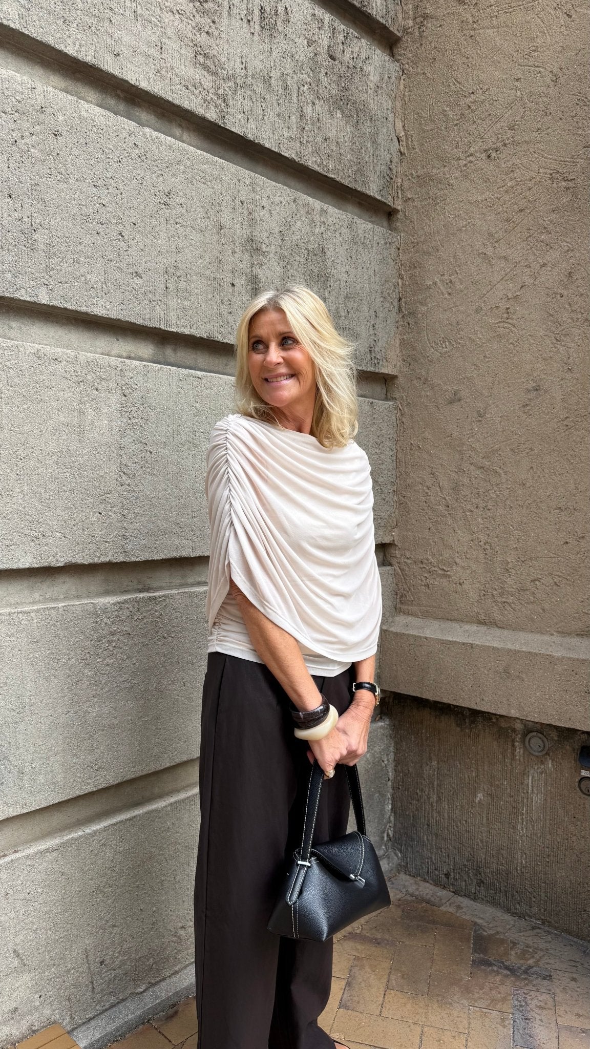 En blond kvinde står ved en betonvæg iført en offwhite Luxzuz Queenie-bluse og mørke bukser og holder en sort taske.