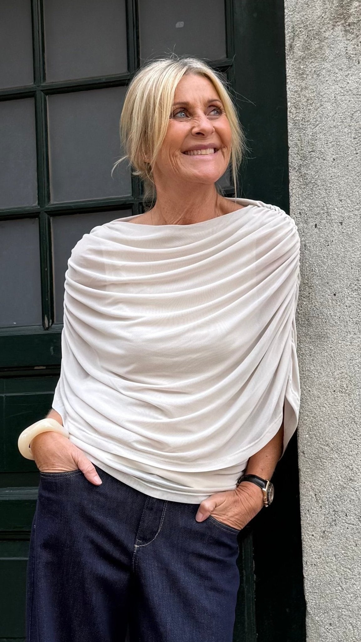 En kvinde med blondt hår har en offwhite Luxzuz Queenie-bluse og mørke bukser på og smiler ved en grøn dør.