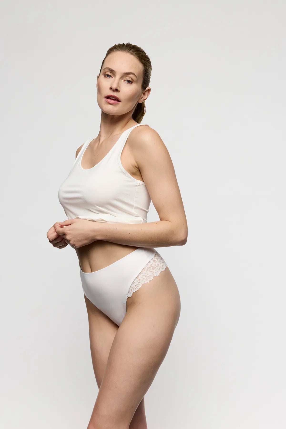 En kvinde i en hvid tanktop og Marie Jo Soft Studio Off White 154739-undertøj står foran en ensfarvet lys baggrund.
