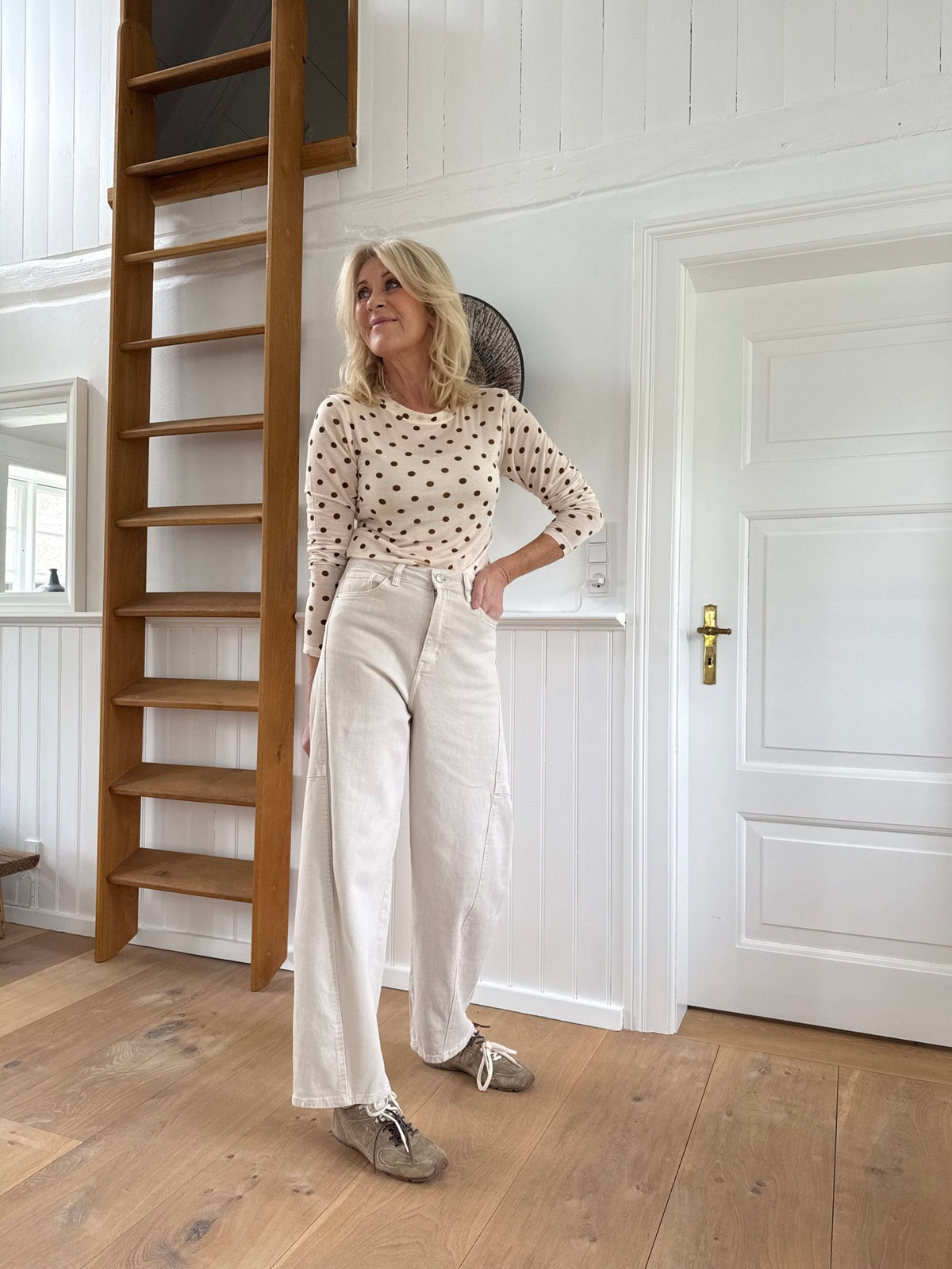 En kvinde står inde i en bluse med prikker og bukser fra Marta du Château – Miltus Beige ved siden af en træstige.