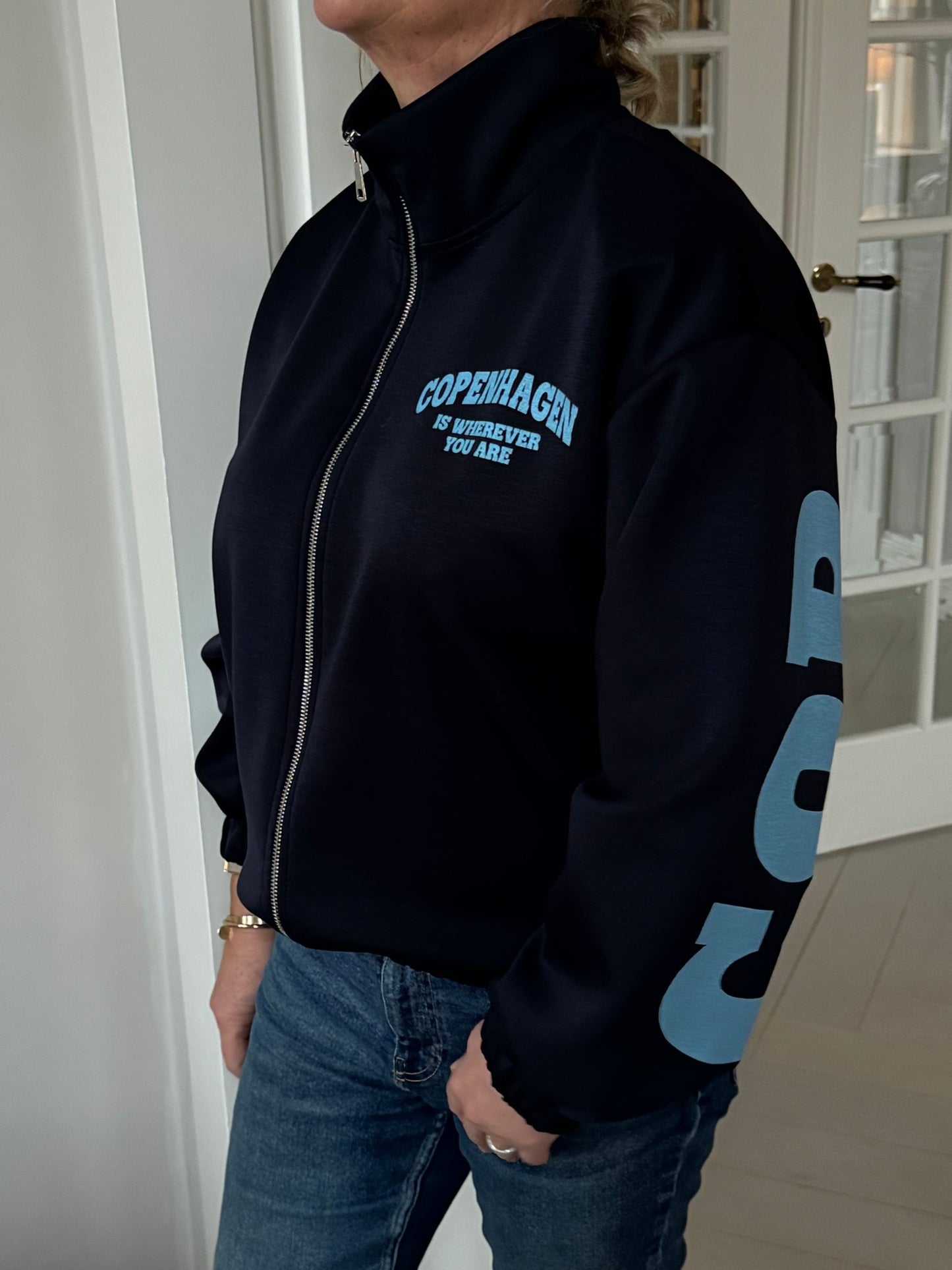 Personen har en Marta du Château Nete Mørkeblå sweatshirt på samt en mørk jakke med lynlås og blå bogstaver på ærmerne.