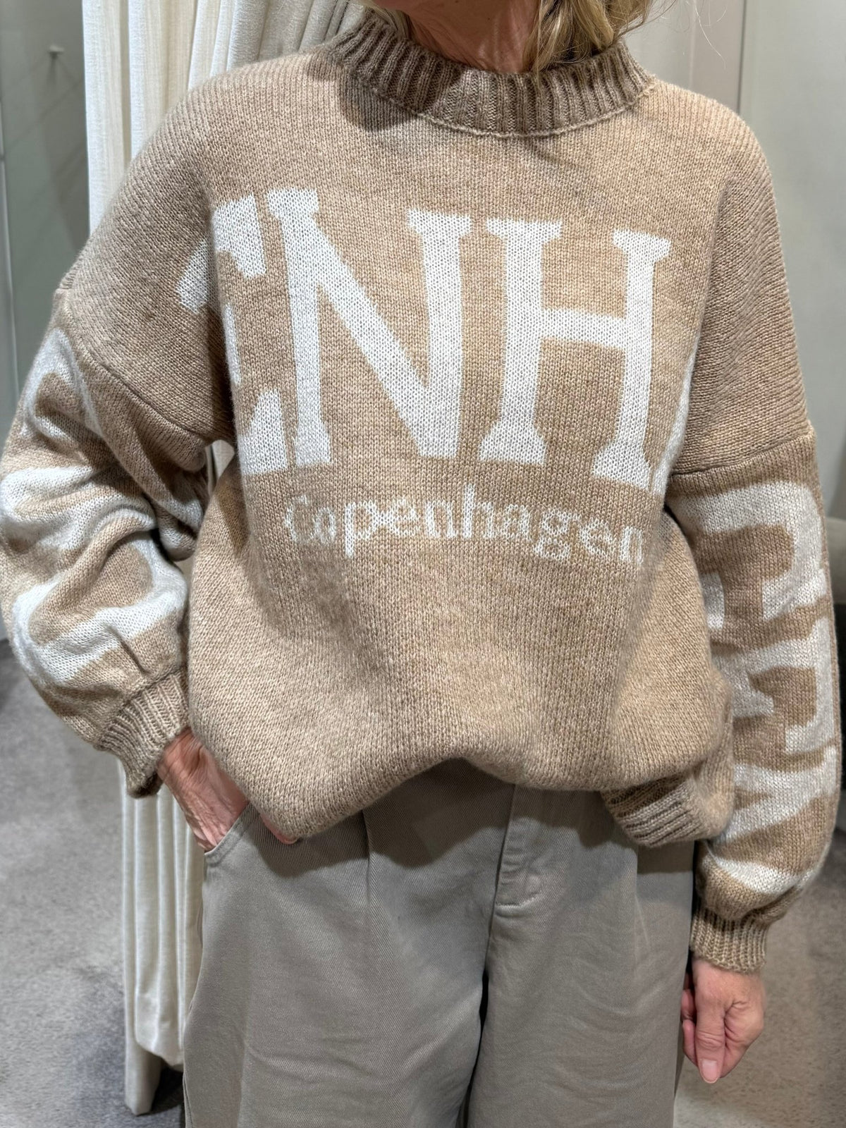 Person iført en Marta du Château Aria Sand-sweater med teksten »CNH copenhagen«, med hånden i lommen.