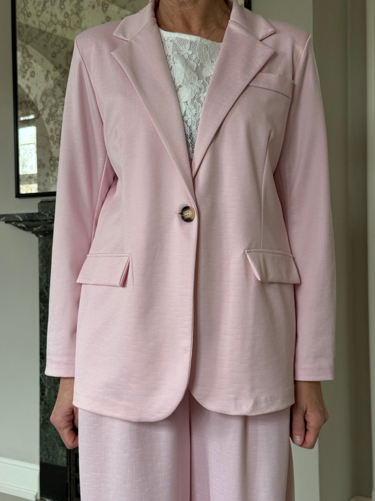 Modellen bærer Marta du Château’s 154405 Anela Lyserød blazer-buksedragt sammen med en hvid bluse i blonder, mens hun er indendørs.