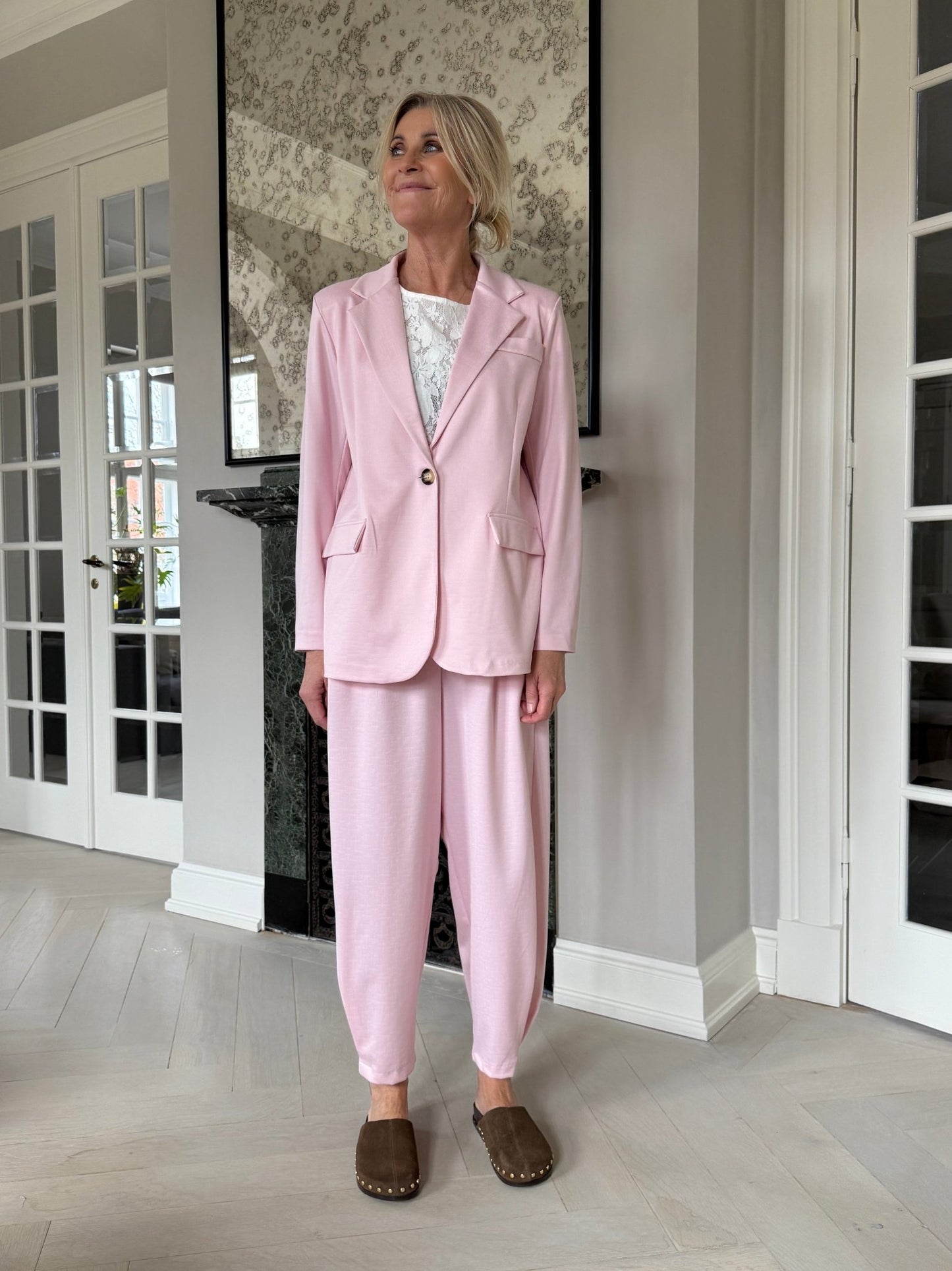 En kvinde iført en Anela Lyserød-blazer fra Marta du Château og en hvid top smiler indenfor, mens de brune sko giver et elegant præg.