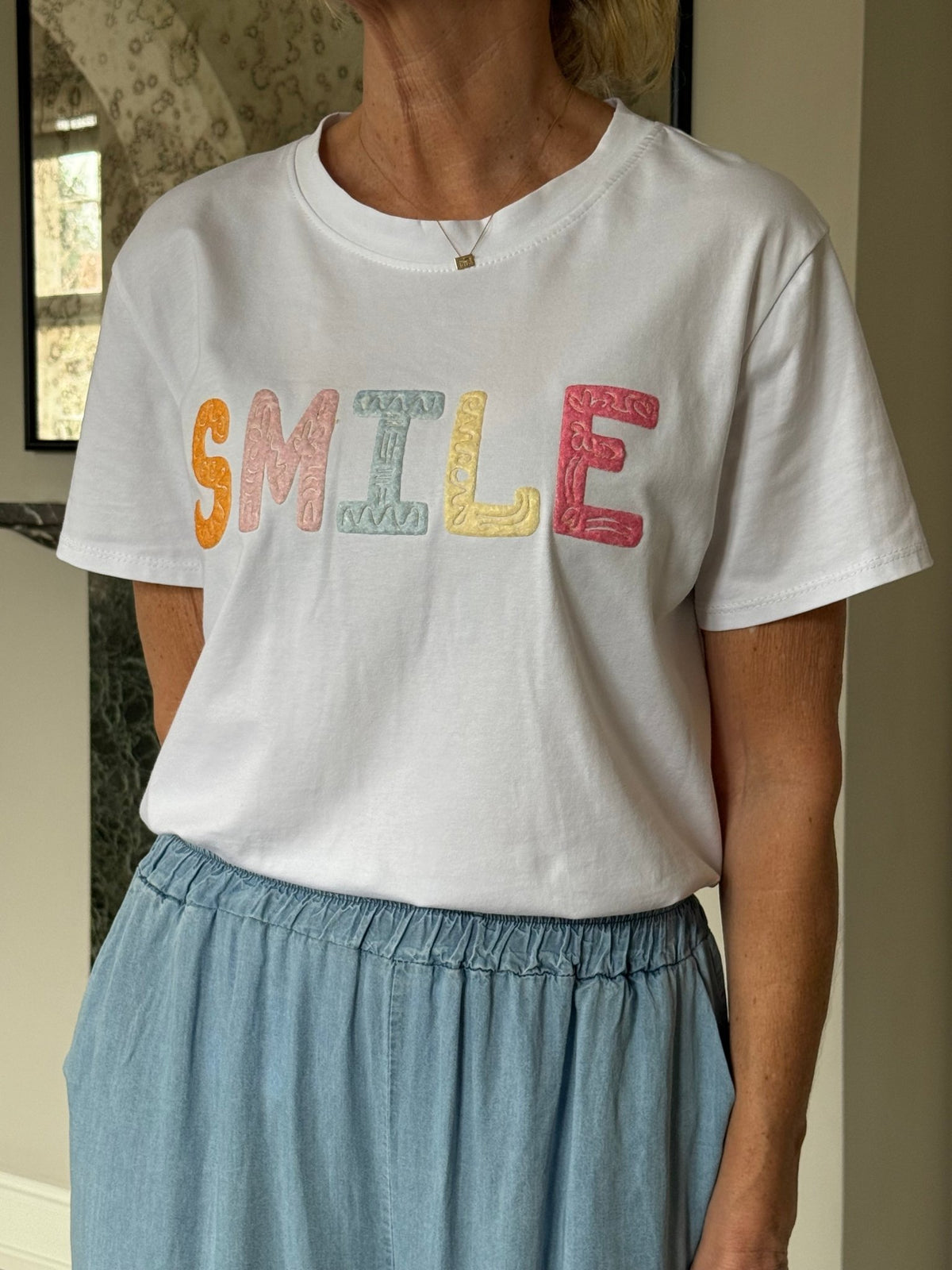 En person iført en hvid T-shirt fra Marta du Château Vespera og en lyseblå nederdel, der står indendørs.