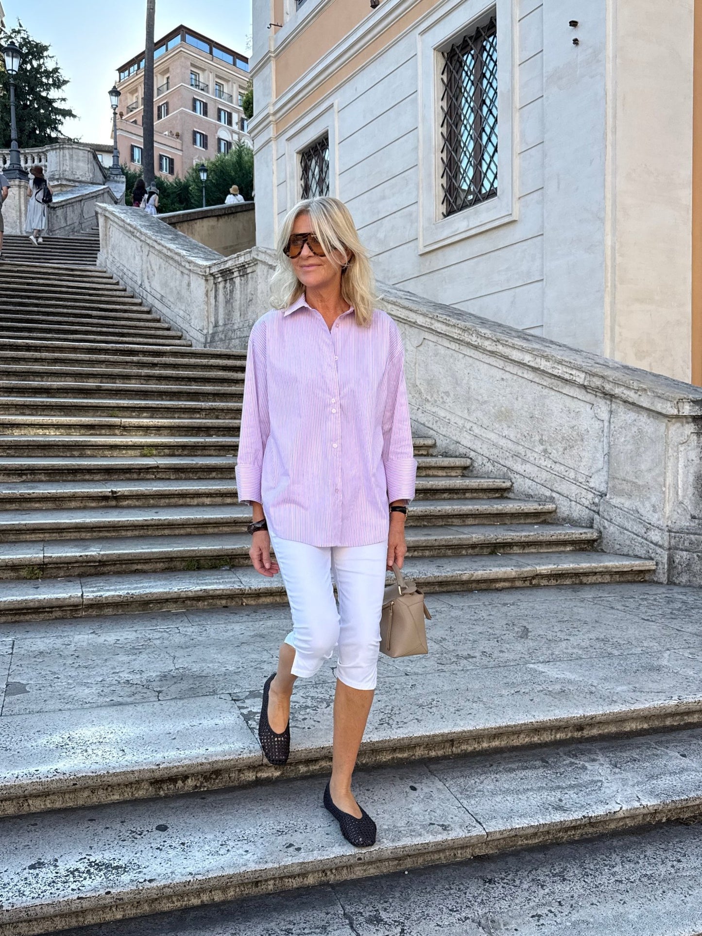 En kvinde i en lyserød bluse og bukser fra Marta du Château Vivianne Hvid går ned ad en stentrappe med en beige taske i hånden.