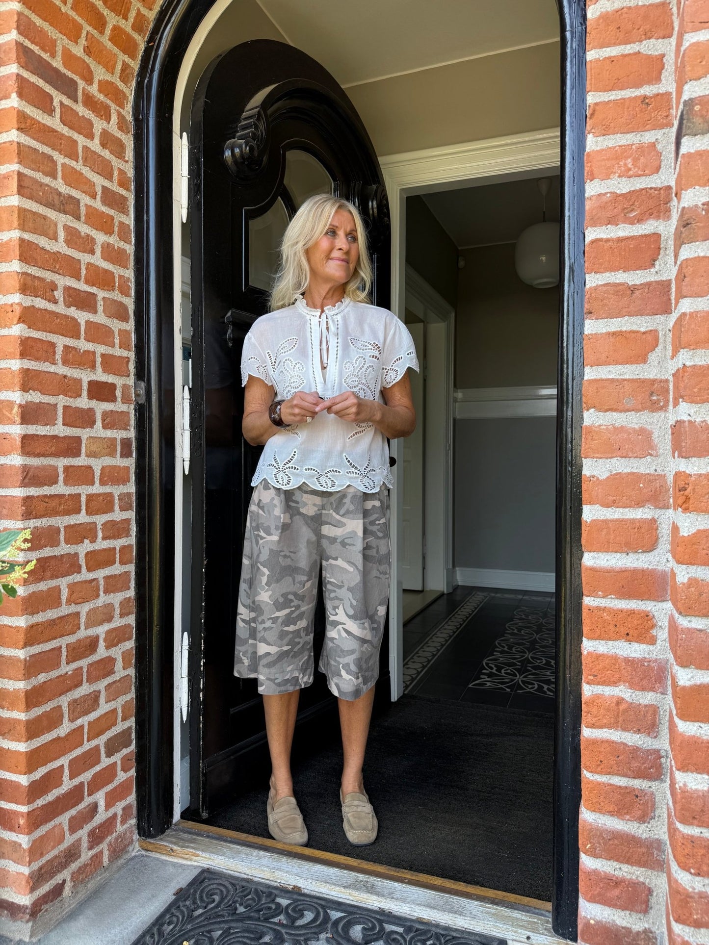 En kvinde i døråbningen har Marta du Château’s Zuri Army-shorts i grøn og en hvid bluse på, med en murstensvæg i baggrunden.