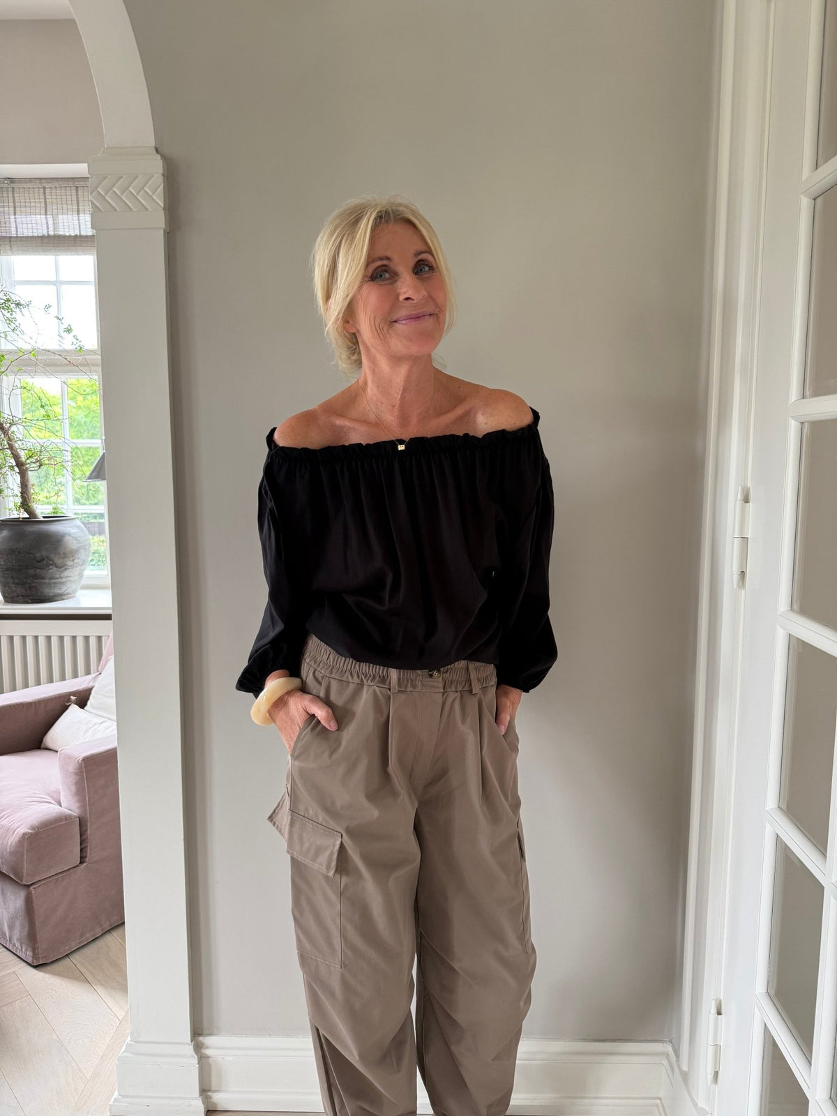 En blond kvinde iført en bluse fra Marta du Château i farven »Juniper Sort« og beige cargobukser står indendørs med hænderne i lommerne.