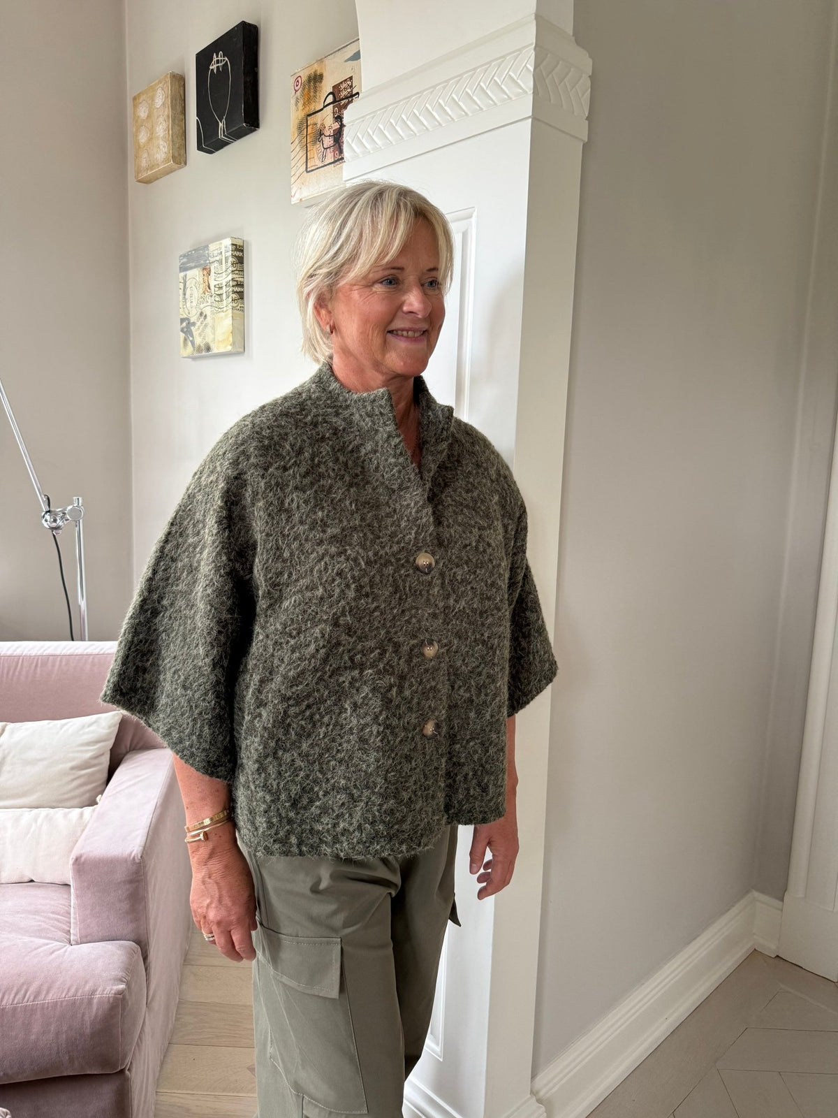 En kvinde sidder indenfor iført en grøn jakke fra Marta du Château Riviera Army ved siden af en lyserød sofa og et vægmaleri.