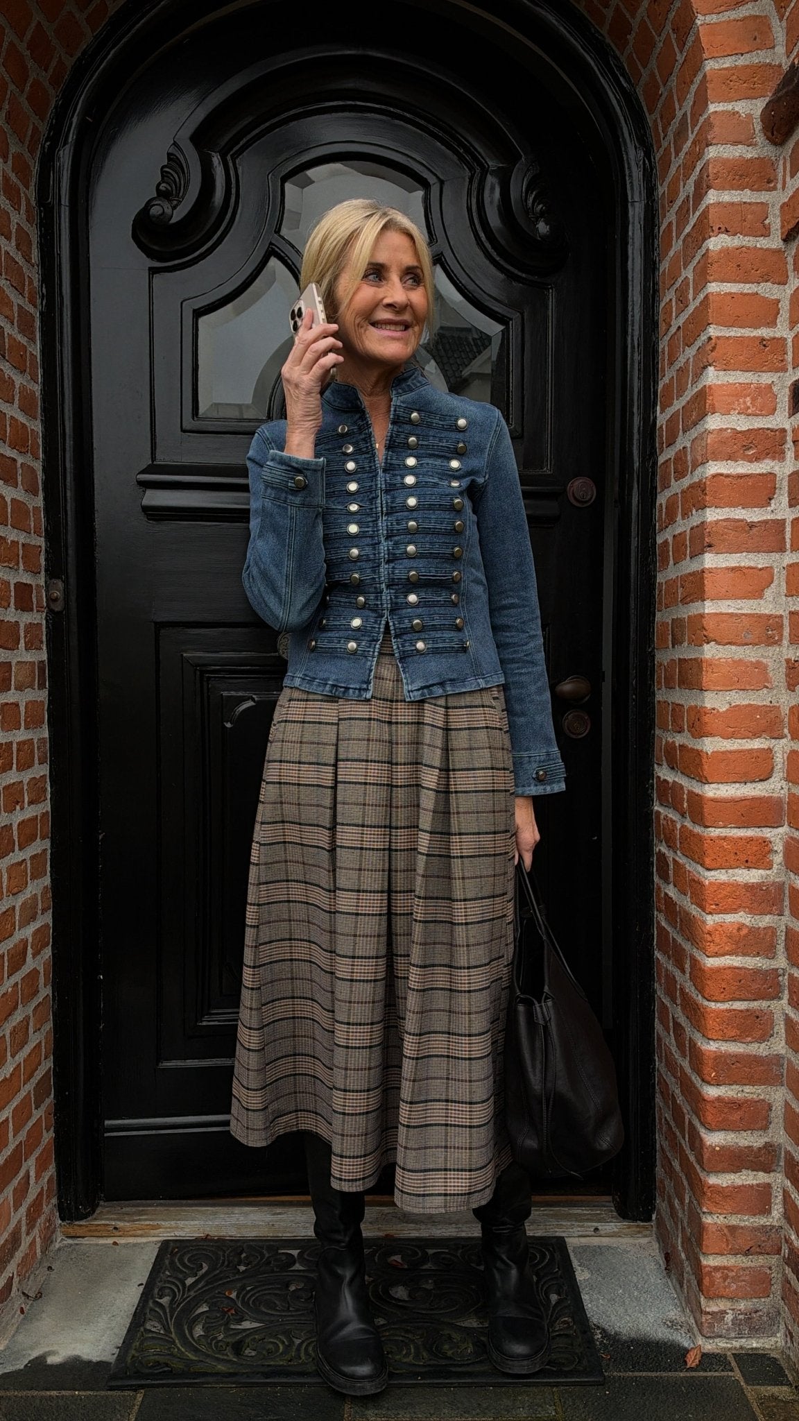 En kvinde iført en 156220 | Marta du Château - MDCAmsonia Ternet-jakke og et ternet midi-nederdel taler i telefon ved en sort dør.