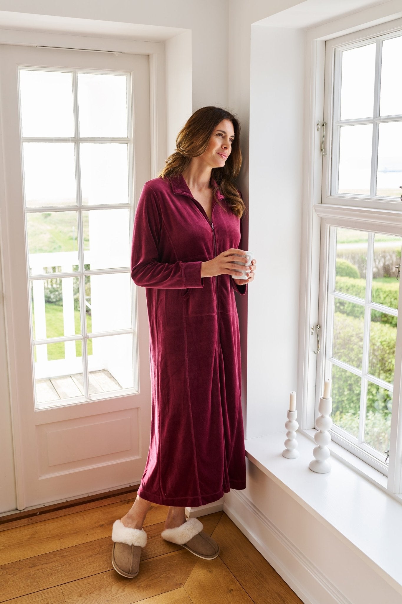 En kvinde i Missyas morgenkåbe i farven »Alice Bordeaux« står ved vinduet og nyder den hyggelige loungewear-stil.