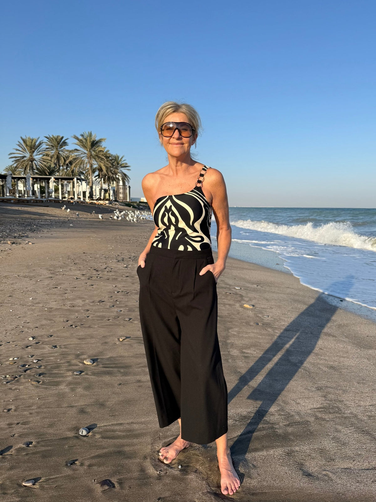 En barfodet kvinde med kort gråt hår har Missya – Capri Sort mønstret på stranden, og solbrillerne giver et elegant præg.